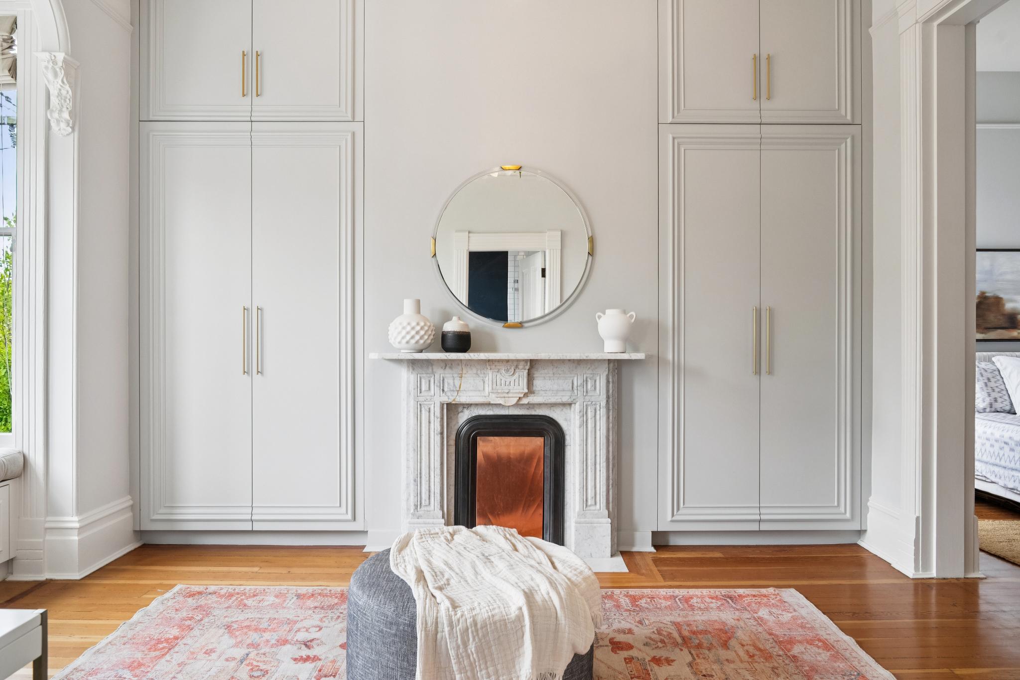 This elegant bedroom features a sophisticated symmetrical design with custom floor-to-ceiling built-in cabinetry flanking a classic marble fireplace. A circular mirror hangs above the mantel, reflecting the room's refined aesthetic, while a plush grey ottoman with a throw blanket sits atop a patterned area rug. The space exudes a serene and luxurious atmosphere, highlighted by high ceilings and polished hardwood flooring.