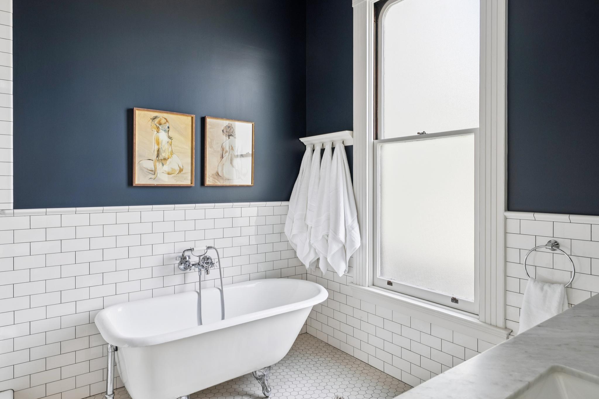 This elegant primary bathroom features a classic clawfoot tub set against a striking navy blue wall and white subway tile wainscoting. A tall, frosted window allows for soft, diffused natural light, while framed artwork and a towel rack add a touch of sophisticated charm. The space is finished with a hexagonal tile floor, creating a timeless and serene atmosphere.