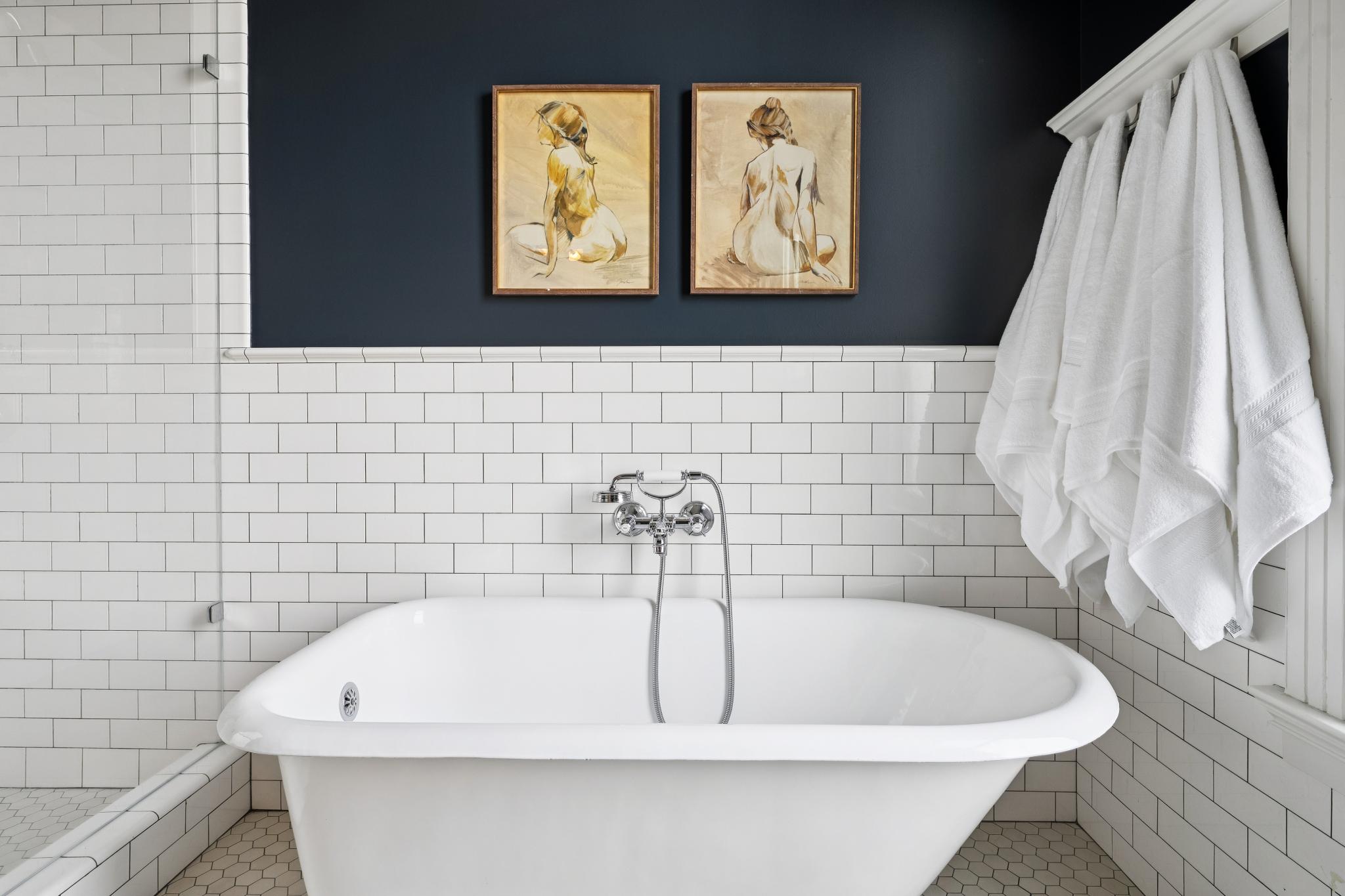 This elegant bathroom features a classic white freestanding soaking tub centered against a wall of white subway tile and a striking dark navy accent wall. Two framed figure sketches hang symmetrically above the tub, while plush white towels are neatly hung on a rack to the right. The space combines a clean, modern aesthetic with traditional charm, highlighted by polished chrome fixtures and hexagonal floor tiles.