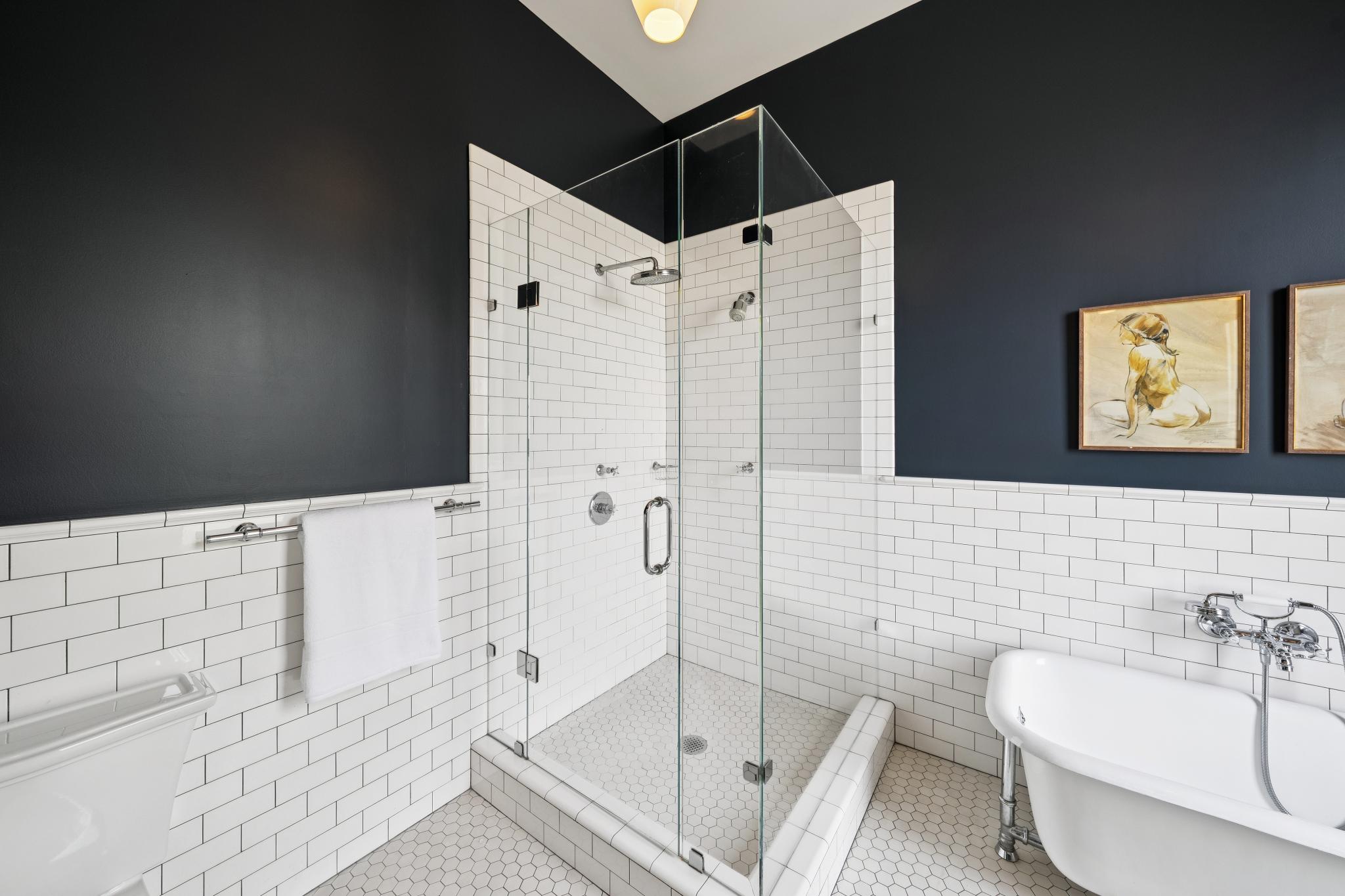 This elegant primary bathroom features a classic black-and-white color palette with subway tile wainscoting and a freestanding clawfoot tub. A glass-enclosed walk-in shower with hexagonal floor tiles serves as a central focal point, complemented by dark charcoal walls and framed artwork. The space exudes a sophisticated, timeless aesthetic that blends vintage charm with modern luxury.