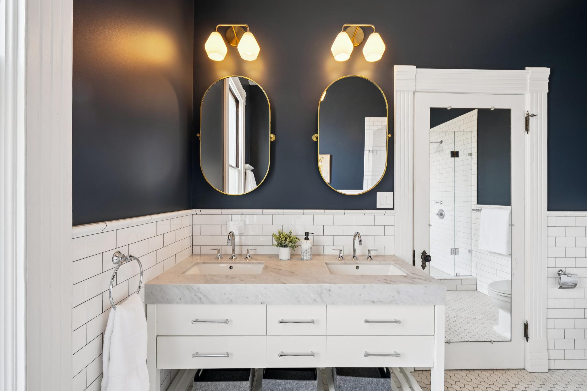 This elegant primary bathroom features a double vanity with a marble countertop, white cabinetry, and dual oval mirrors accented by gold-toned wall sconces. The space is defined by a striking contrast between dark navy walls and classic white subway tile wainscoting, creating a sophisticated, modern aesthetic. A doorway with a full-length mirror leads into a bright, tiled shower area, enhancing the sense of depth and luxury.