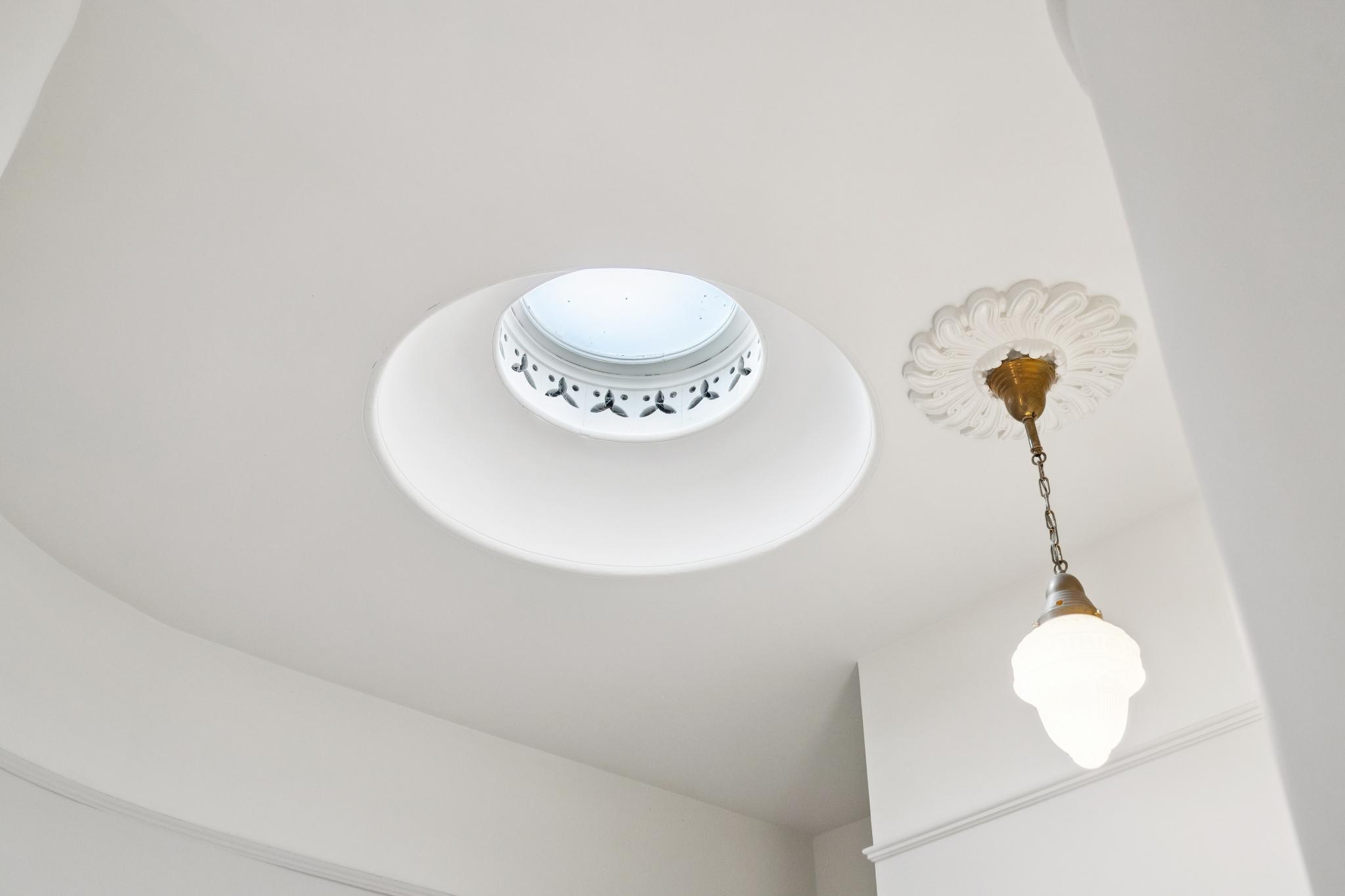 This image highlights a sophisticated architectural detail featuring a circular skylight recessed into a smooth, white ceiling. Adjacent to the skylight is an elegant, vintage-style pendant light fixture suspended from an ornate, decorative ceiling medallion. The composition emphasizes the clean lines and classic design elements, creating a bright and refined atmosphere.