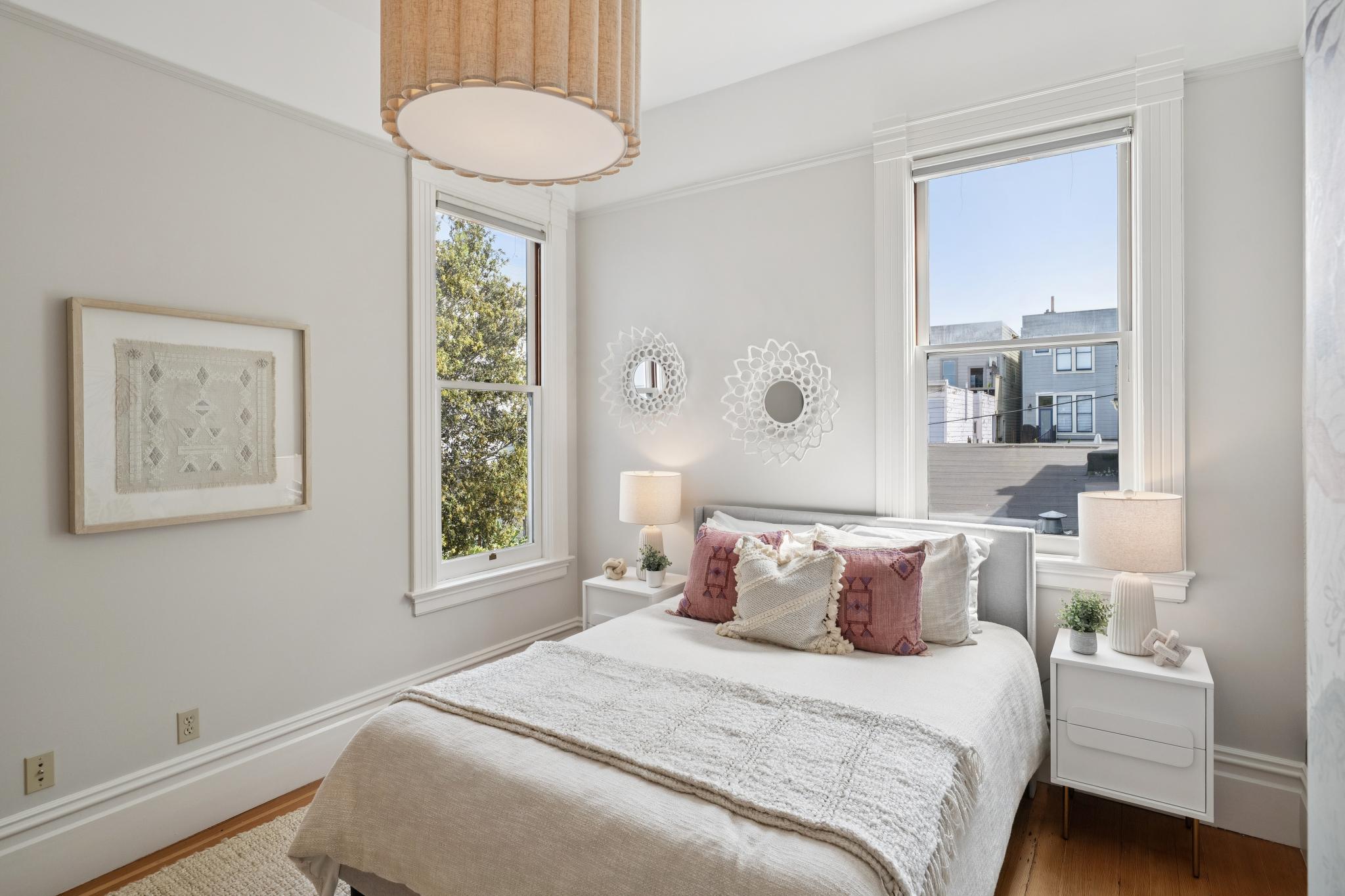 This bright and airy bedroom features a neutral color palette with soft textures, centered around a cozy bed adorned with decorative pillows. Two white nightstands with matching lamps flank the bed, while unique circular wall art and a large drum pendant light add a touch of modern elegance. The room is framed by two windows that allow ample natural light to fill the space, creating a serene and inviting atmosphere.