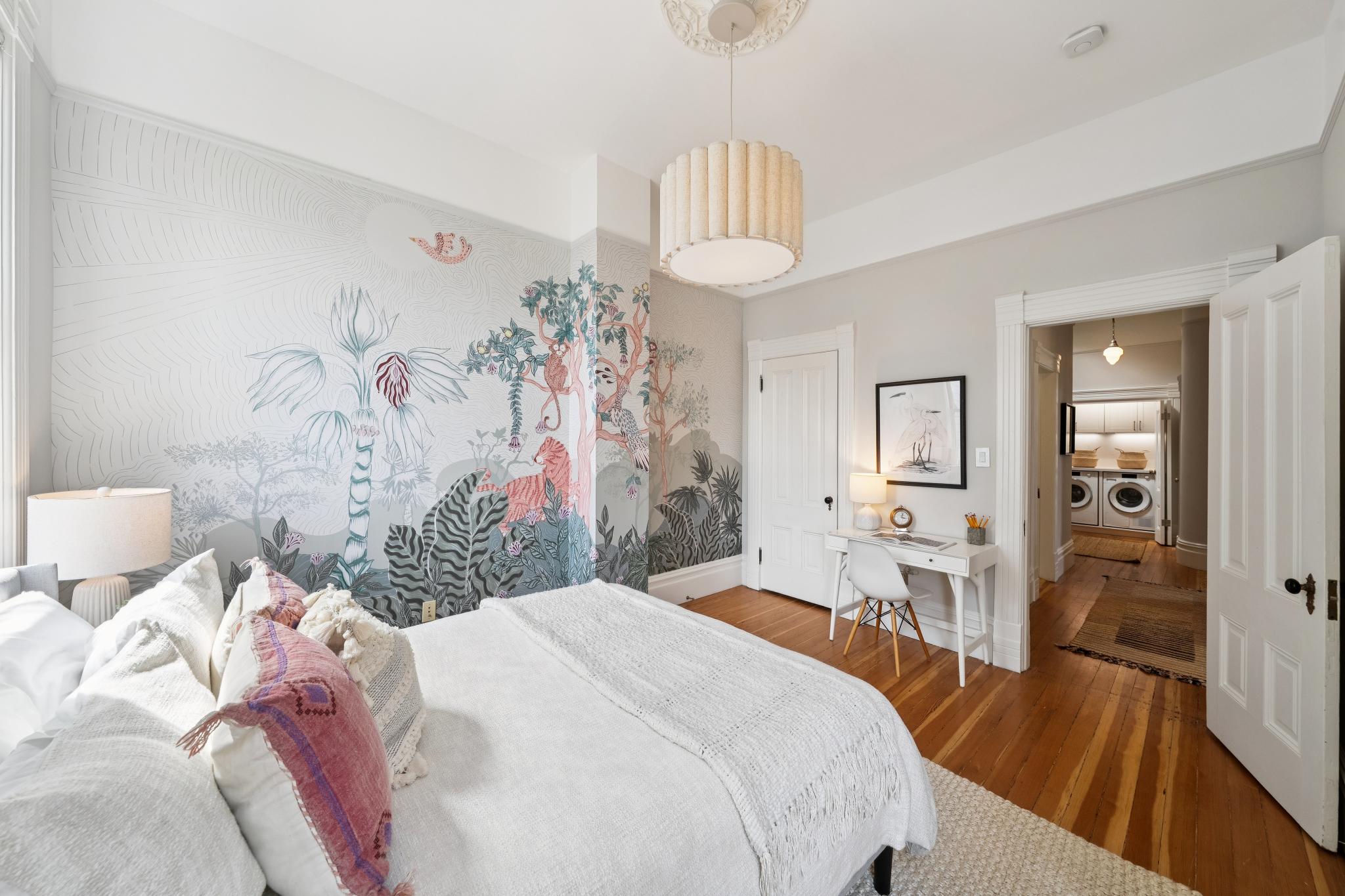 This bright and inviting bedroom features a striking jungle-themed mural on the accent wall, complemented by a neutral color palette and warm hardwood flooring. The room is furnished with a comfortable bed, a sleek white desk, and a stylish pendant light, creating a serene and artistic atmosphere. The perspective looks from the foot of the bed toward a doorway that leads into a laundry area, highlighting the home's functional layout.