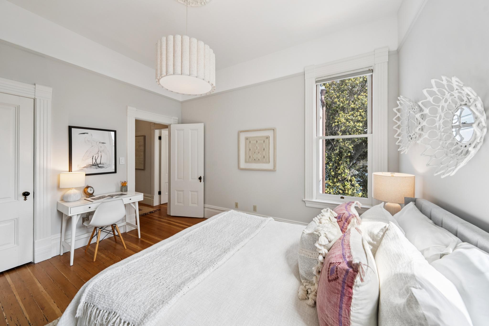This bright and airy primary bedroom features a neutral color palette, hardwood floors, and a large window that invites natural light. The room is furnished with a comfortable bed adorned with textured pillows, a sleek white desk setup, and a unique decorative wall mirror, creating a serene and functional living space. The perspective is from the foot of the bed, capturing the cohesive design and inviting atmosphere of the room.