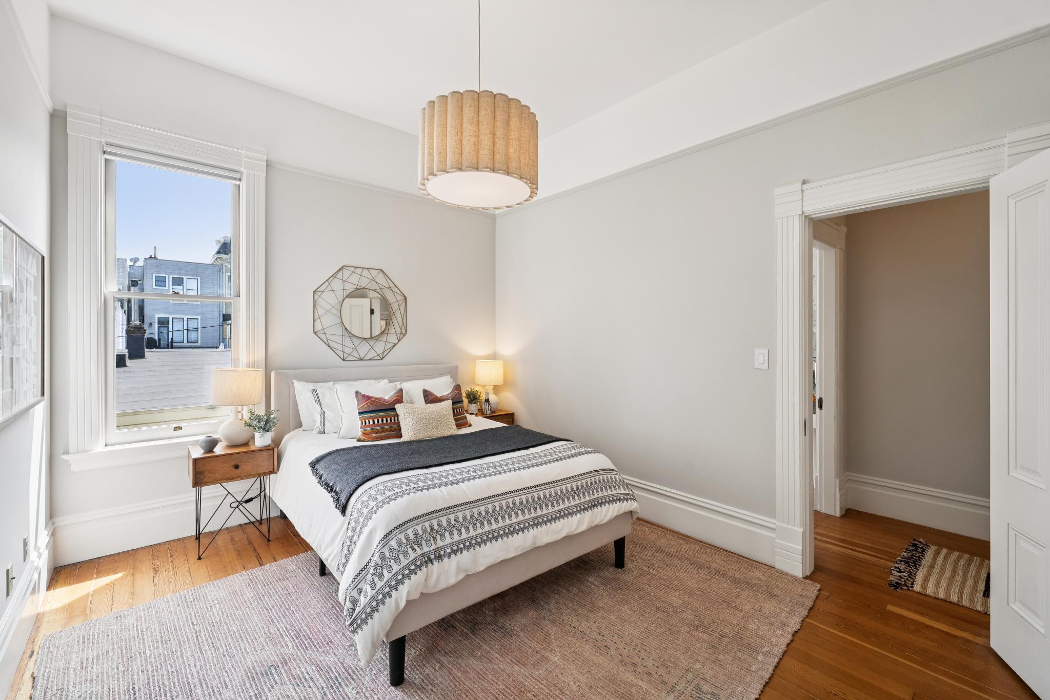 This bright and inviting bedroom features a neutral color palette with warm hardwood floors and classic architectural trim. The room is centered around a queen-sized bed adorned with patterned textiles, flanked by two minimalist wooden nightstands and matching lamps. A large window allows for natural light, while a unique geometric wall mirror adds a modern touch to the space.