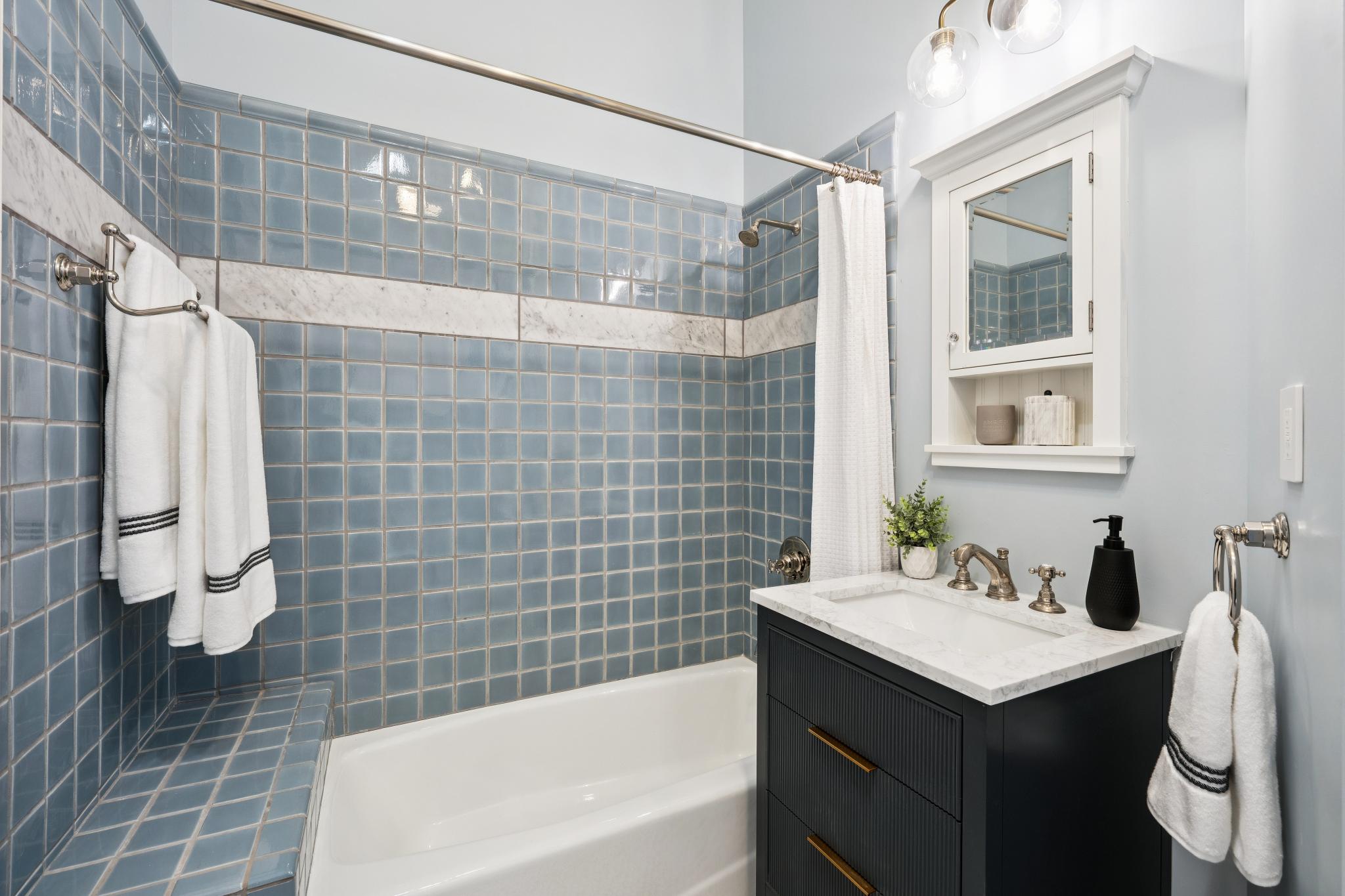 This charming bathroom features a classic blue-tiled shower surround with a marble accent stripe and a matching tiled bench. A sleek, dark-toned vanity with a white marble countertop and gold hardware provides a modern contrast to the vintage-inspired wall tiles. The space is completed by a white medicine cabinet, a matching towel ring, and soft, warm lighting, creating a clean and inviting atmosphere.