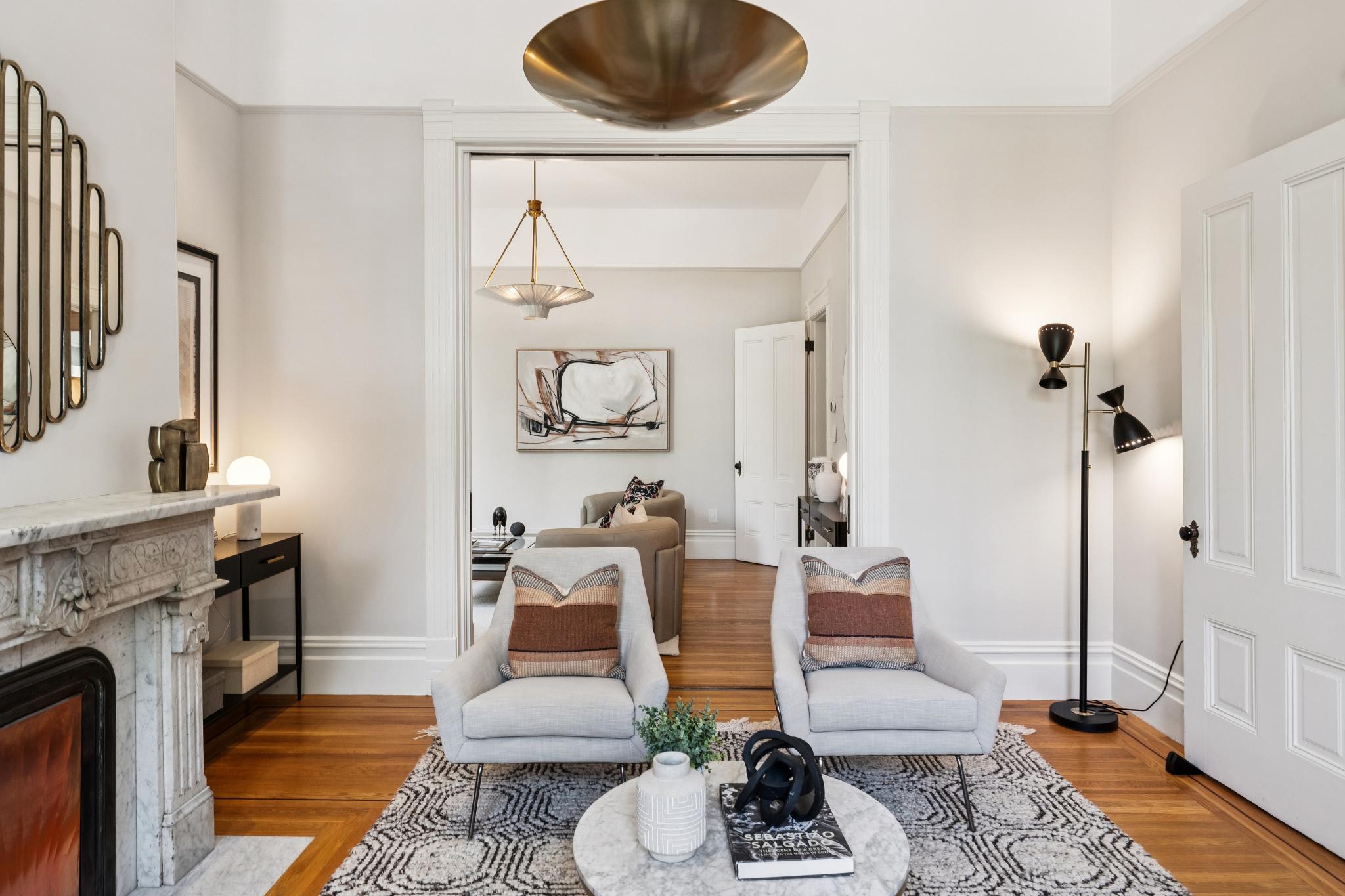 This elegant living room features a symmetrical arrangement of two light gray armchairs facing a marble-topped coffee table, set upon a textured geometric rug. A classic marble fireplace mantel anchors the left side, while a wide doorway leads into an adjacent sitting area, creating a sense of depth and flow. The space is defined by high ceilings, warm hardwood floors, and a sophisticated blend of modern lighting fixtures and curated decor.