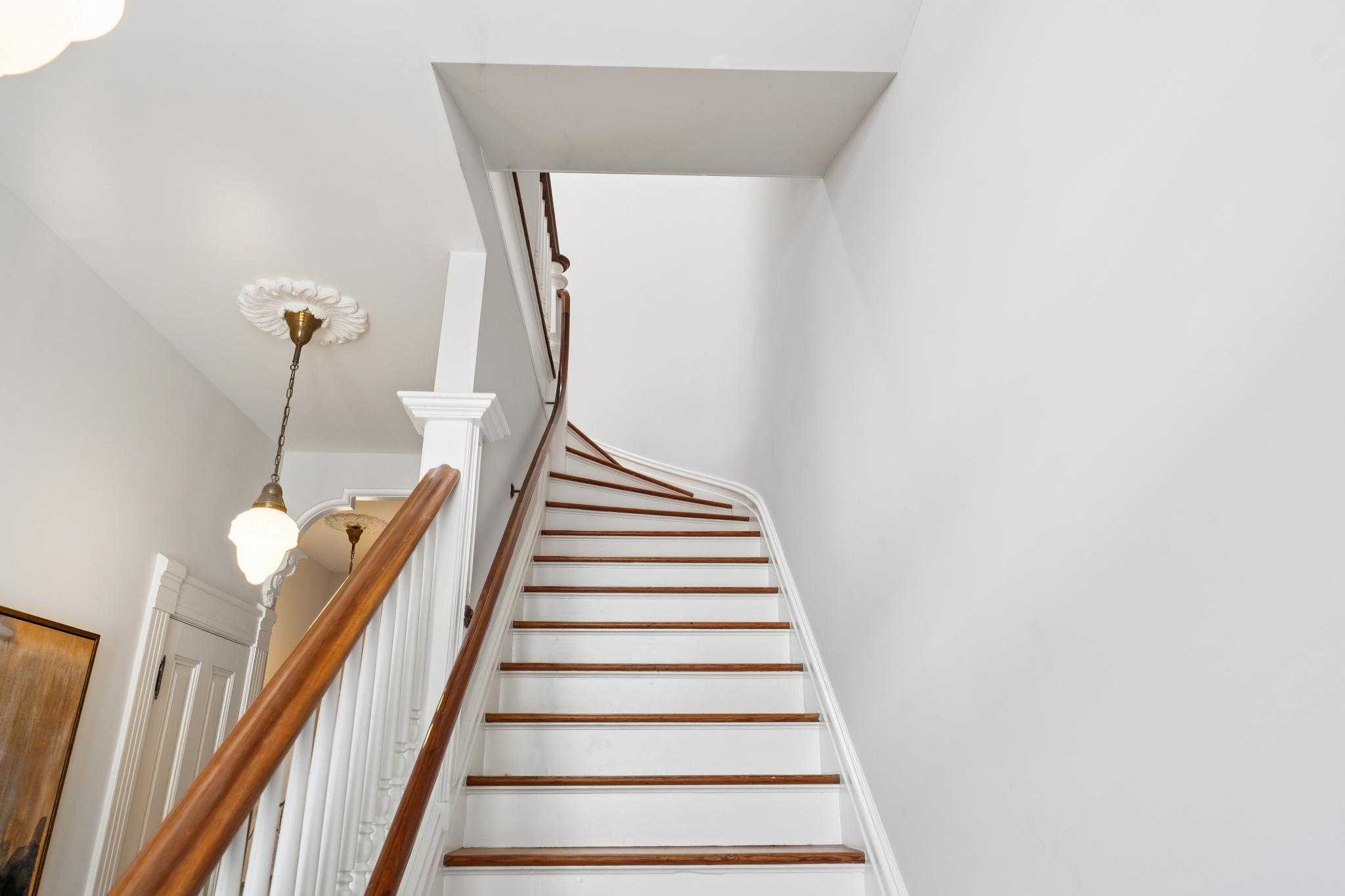 This image captures a classic, elegant staircase featuring white risers and rich, stained wood treads that curve gracefully upward. A polished wooden handrail supported by white balusters leads the eye toward the upper level, while a vintage-style pendant light fixture hangs from a decorative ceiling medallion. The perspective is from the bottom of the stairs, emphasizing the architectural charm and clean, bright aesthetic of the home's entryway.