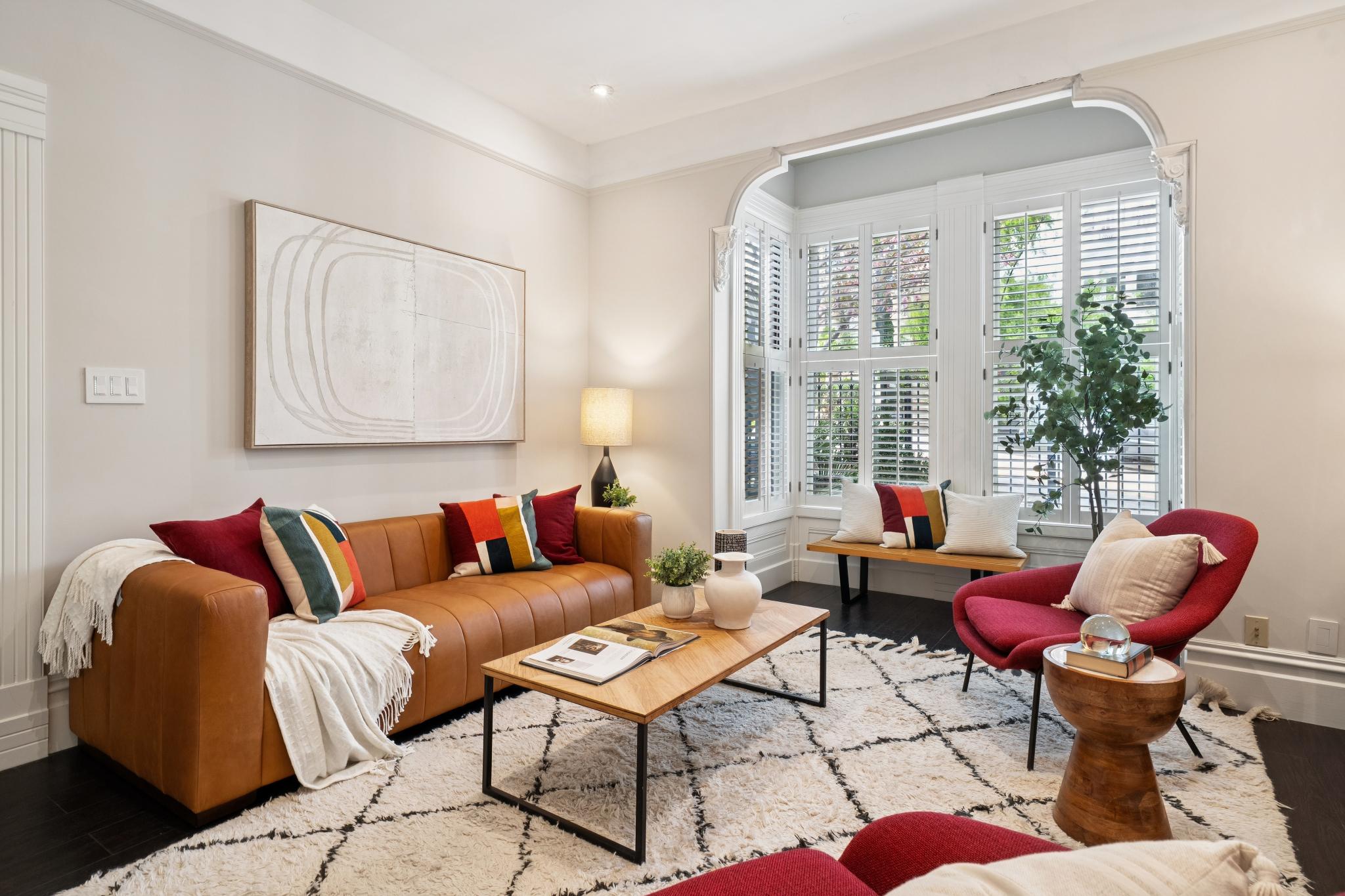 This inviting living room features a warm, modern aesthetic anchored by a rich tan leather sofa and vibrant red accent chairs. A large, textured area rug sits beneath a minimalist wooden coffee table, while a bay window with plantation shutters floods the space with natural light. The room is tastefully decorated with abstract wall art and a potted plant, creating a cozy and sophisticated atmosphere.
