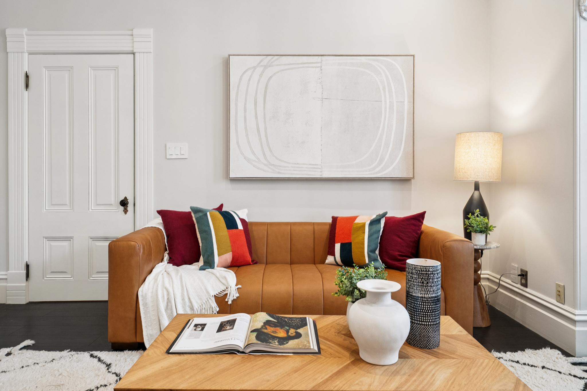 This inviting living room space features a warm, cognac-colored leather sofa accented with vibrant, multi-colored throw pillows and a soft white throw blanket. A light wood coffee table sits in the foreground, topped with an open book and decorative vases, while a large, minimalist abstract art piece hangs centered above the sofa. The room is grounded by dark hardwood floors and a neutral color palette, creating a cozy and sophisticated atmosphere.