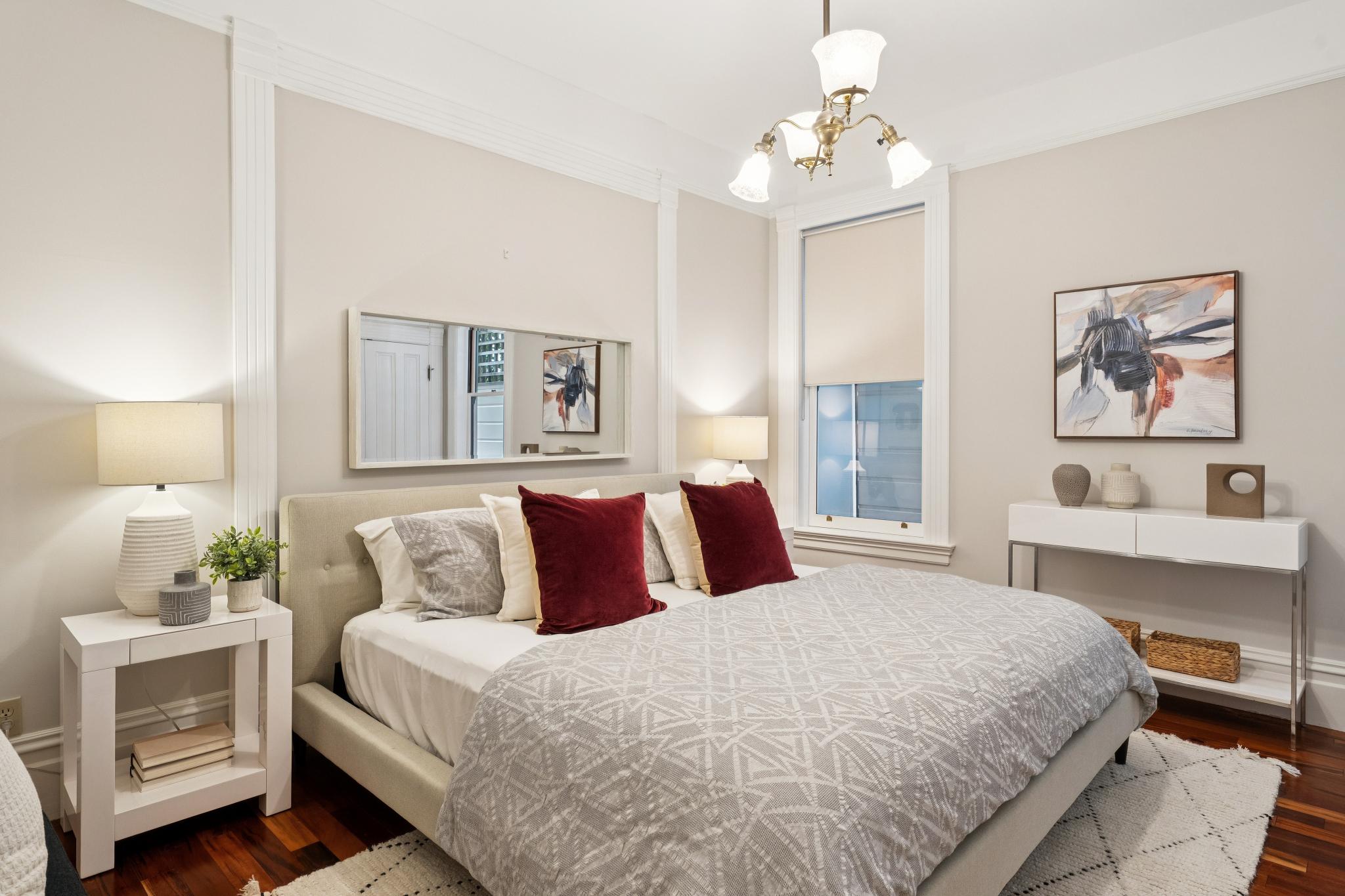 This elegantly staged primary bedroom features a neutral color palette with a plush upholstered bed frame and a patterned grey duvet. The room is accented by deep red throw pillows, a modern chandelier, and a sleek white console table, creating a sophisticated and inviting atmosphere. A large mirror above the bed enhances the sense of space, while the warm wood flooring adds a touch of classic charm.