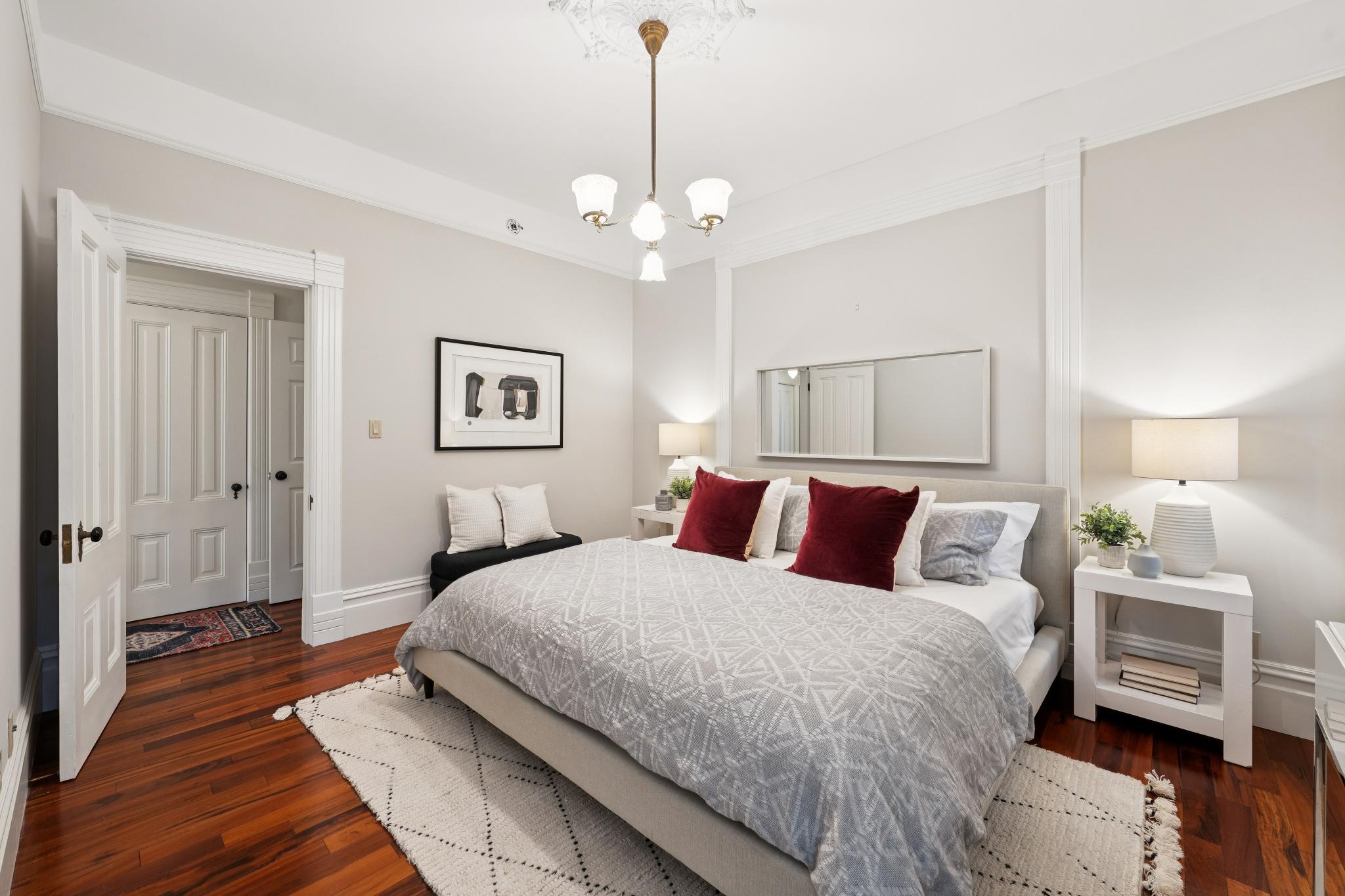 This elegant primary bedroom features rich hardwood flooring, a neutral color palette, and a sophisticated chandelier that adds a touch of classic charm. The room is anchored by a plush bed with a patterned duvet and bold red accent pillows, flanked by matching white nightstands and lamps. A large mirror above the headboard enhances the sense of space, while the open doorway leads to a bright, inviting hallway.