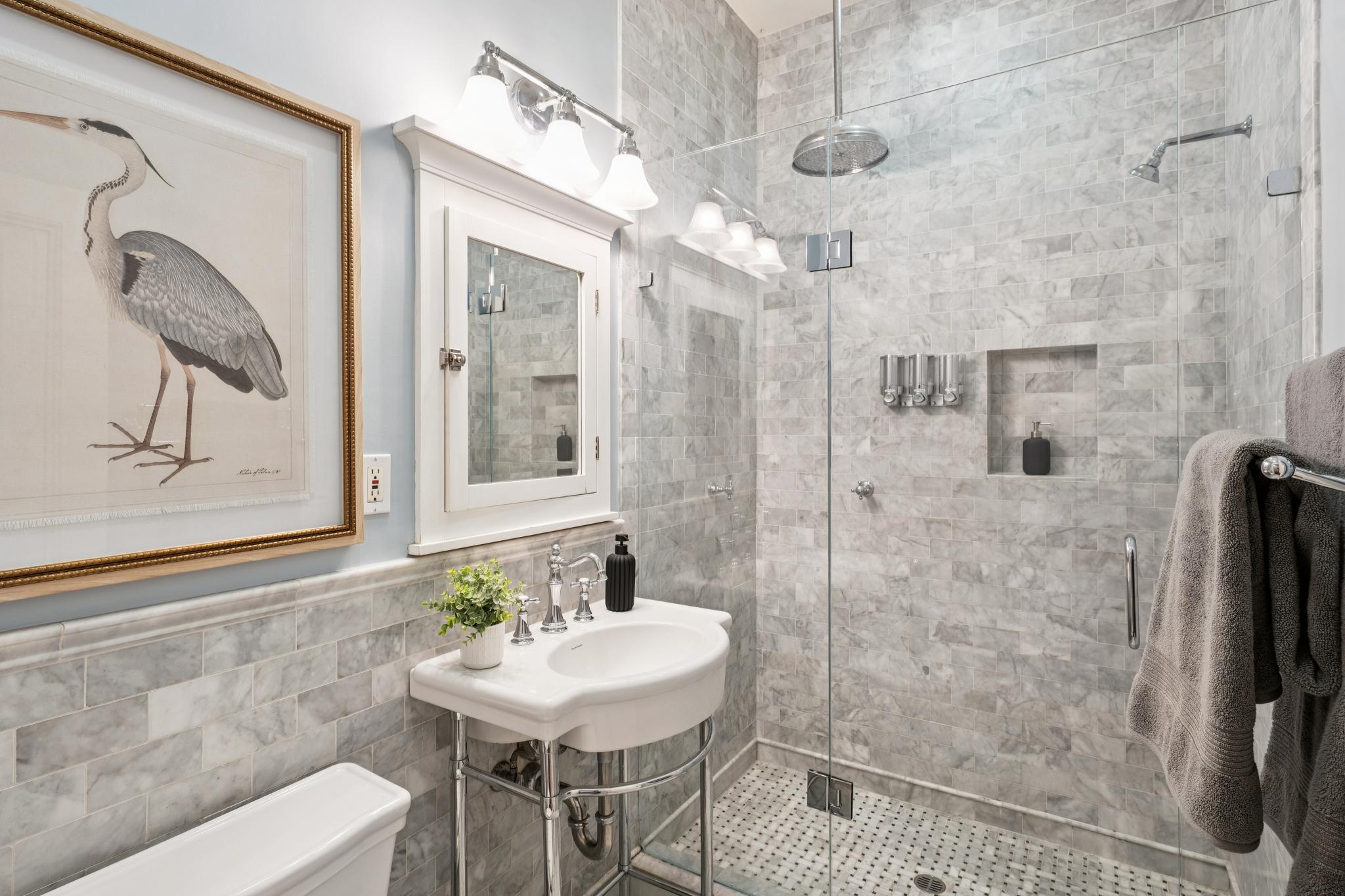This elegant bathroom features a classic pedestal sink, a glass-enclosed walk-in shower with marble subway tile, and a sophisticated heron print framed on the wall. The space is accented by polished chrome fixtures, a medicine cabinet with integrated lighting, and a neutral color palette that creates a clean, timeless aesthetic. The perspective is a medium-wide shot capturing the functional layout and high-end finishes of the room.