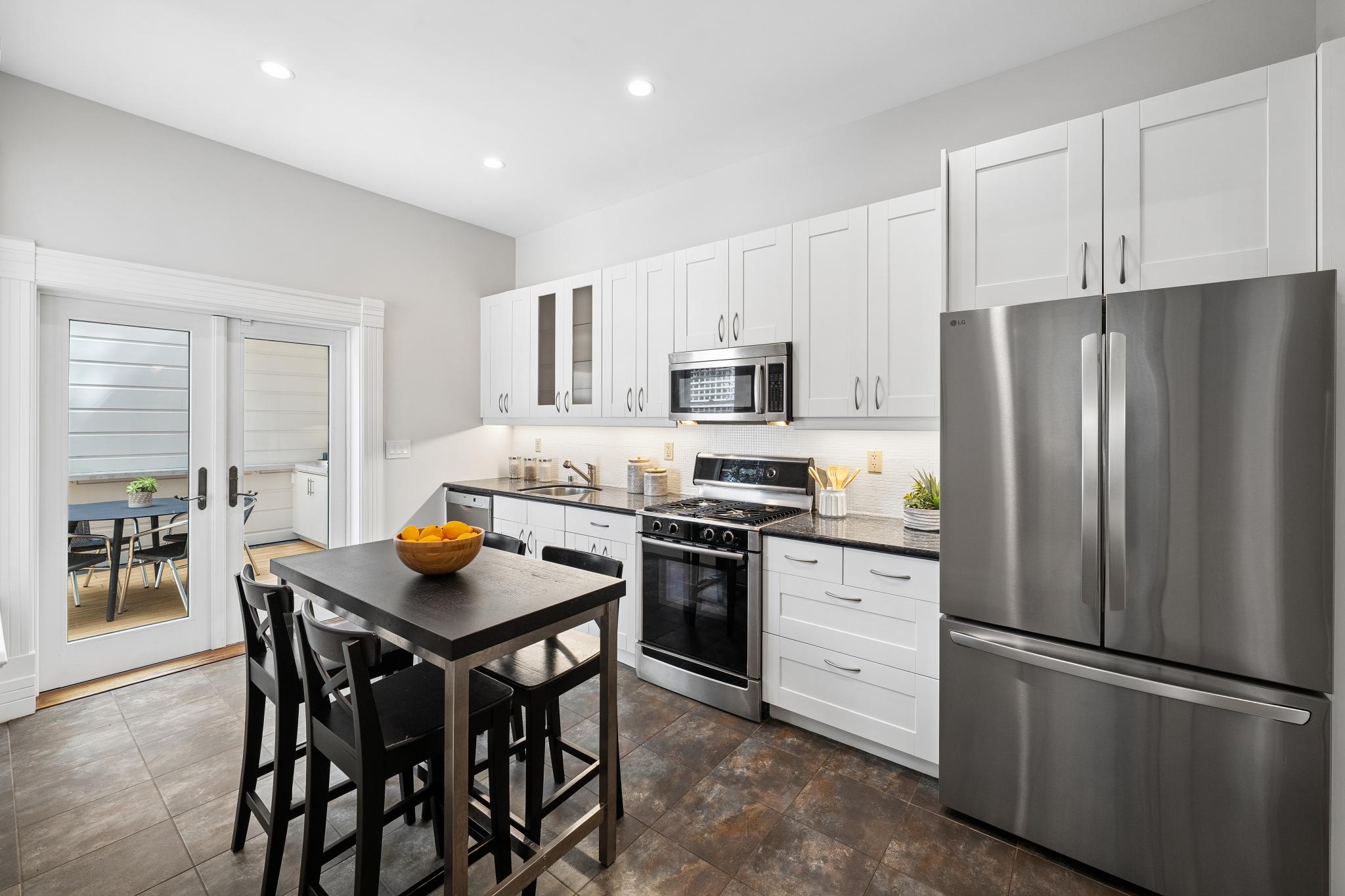 This bright and modern kitchen features crisp white cabinetry paired with dark granite countertops and stainless steel appliances, including a large refrigerator and a gas range. A central dining island with black chairs provides a functional eating space, while glass-paned French doors offer a seamless transition to an outdoor patio area. The room is finished with neutral wall tones and durable tile flooring, creating a clean and inviting atmosphere.