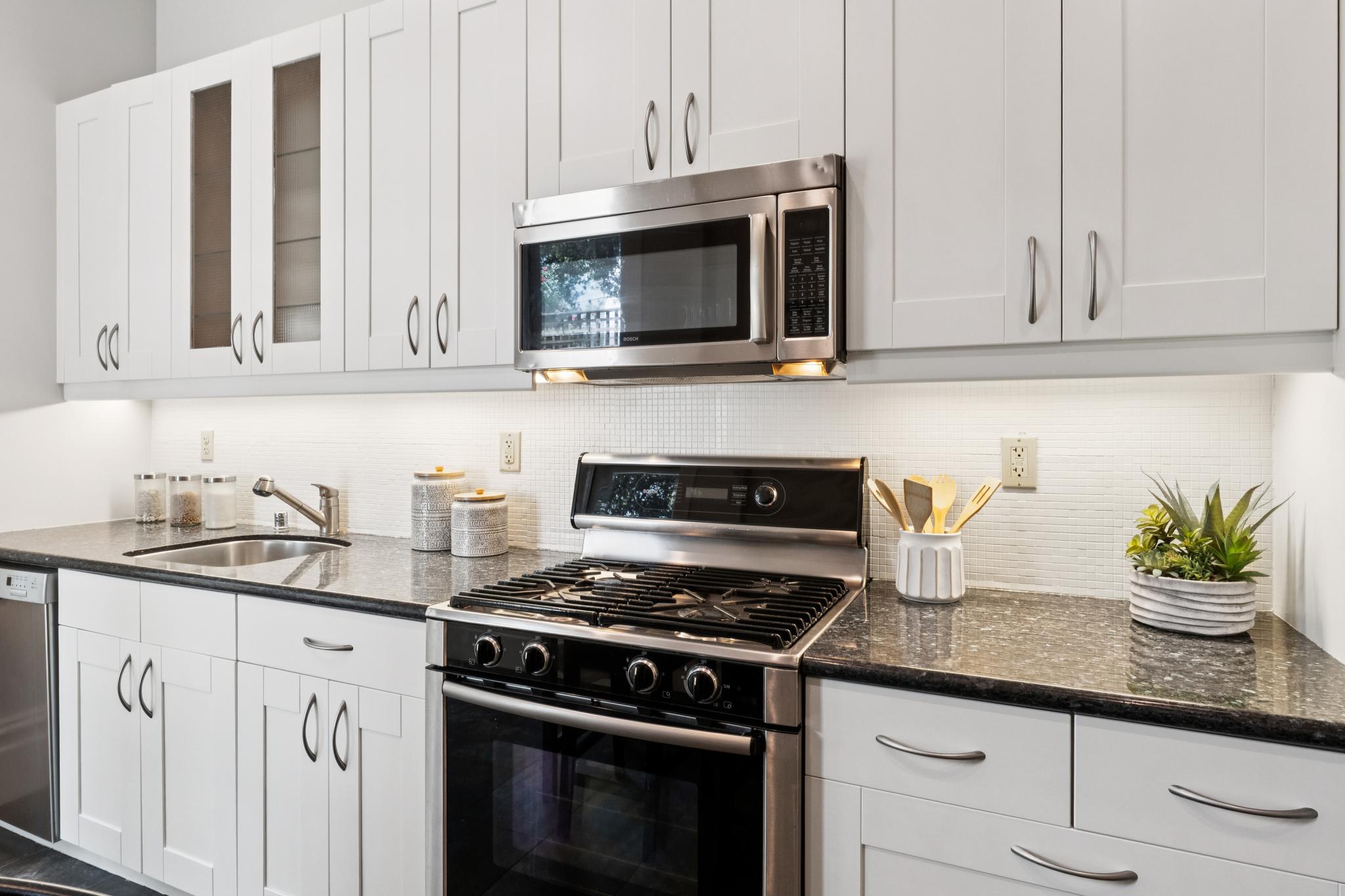 This modern kitchen features crisp white shaker-style cabinetry paired with dark granite countertops and a clean white subway tile backsplash. The space is equipped with stainless steel appliances, including a gas range and an over-the-range microwave, creating a functional and polished culinary environment. A small potted plant and a utensil holder add a touch of warmth to the sleek, contemporary design.