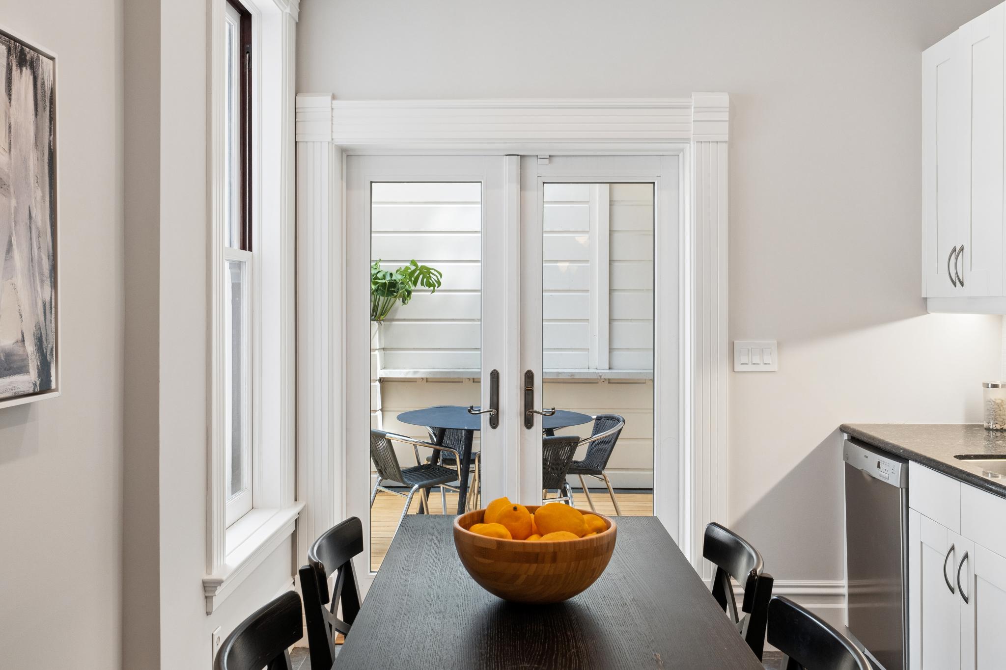 This dining area features a dark wood table centered in the frame, topped with a wooden bowl filled with fresh lemons, and surrounded by black chairs. The space opens through classic white French doors to a small, inviting outdoor deck with a bistro set. To the right, a glimpse of a clean, white-cabinet kitchen adds to the functional and bright atmosphere of this modern home.