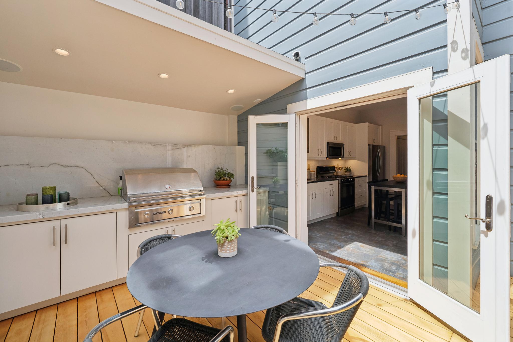 This inviting outdoor patio features a sleek, modern built-in grill station with marble countertops and white cabinetry, seamlessly connecting to an indoor kitchen through wide-open French doors. A dark circular dining table with matching chairs sits on the warm-toned wooden deck, creating a perfect space for al fresco dining. The scene is framed by string lights overhead and a view into the bright, well-appointed interior kitchen, emphasizing a fluid indoor-outdoor living concept.