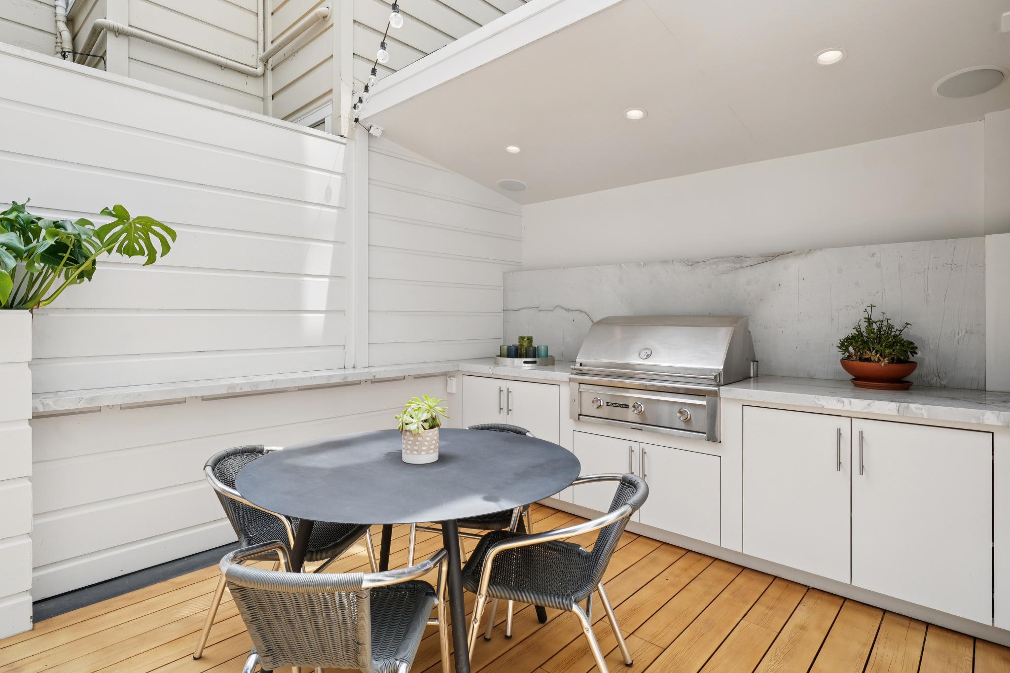 This inviting outdoor patio features a sleek, modern built-in kitchen with a stainless steel grill and white cabinetry set against a marble backsplash. A circular black dining table with matching woven chairs sits on a warm-toned wooden deck, creating a perfect space for al fresco dining. The area is sheltered by a clean, white-paneled structure with recessed lighting, offering a sophisticated and functional outdoor living experience.