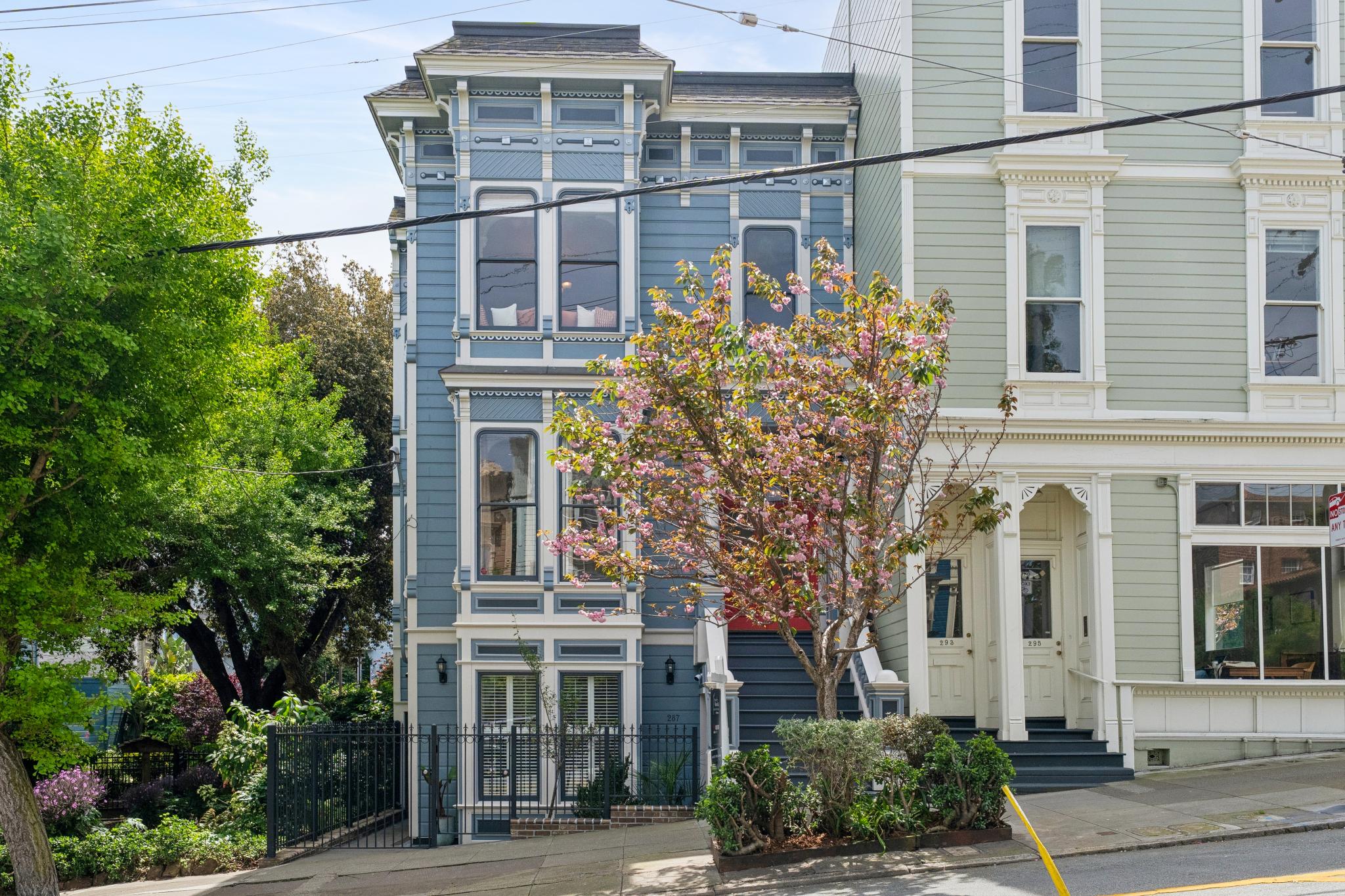 This image captures a charming, multi-story Victorian-style townhouse painted in a soft blue with white trim, showcasing intricate architectural details and a classic bay window design. A vibrant, blooming cherry blossom tree stands in the foreground, adding a touch of natural beauty to the urban streetscape. The perspective is a street-level shot that highlights the home's historic character and its position next to a neighboring light green building.