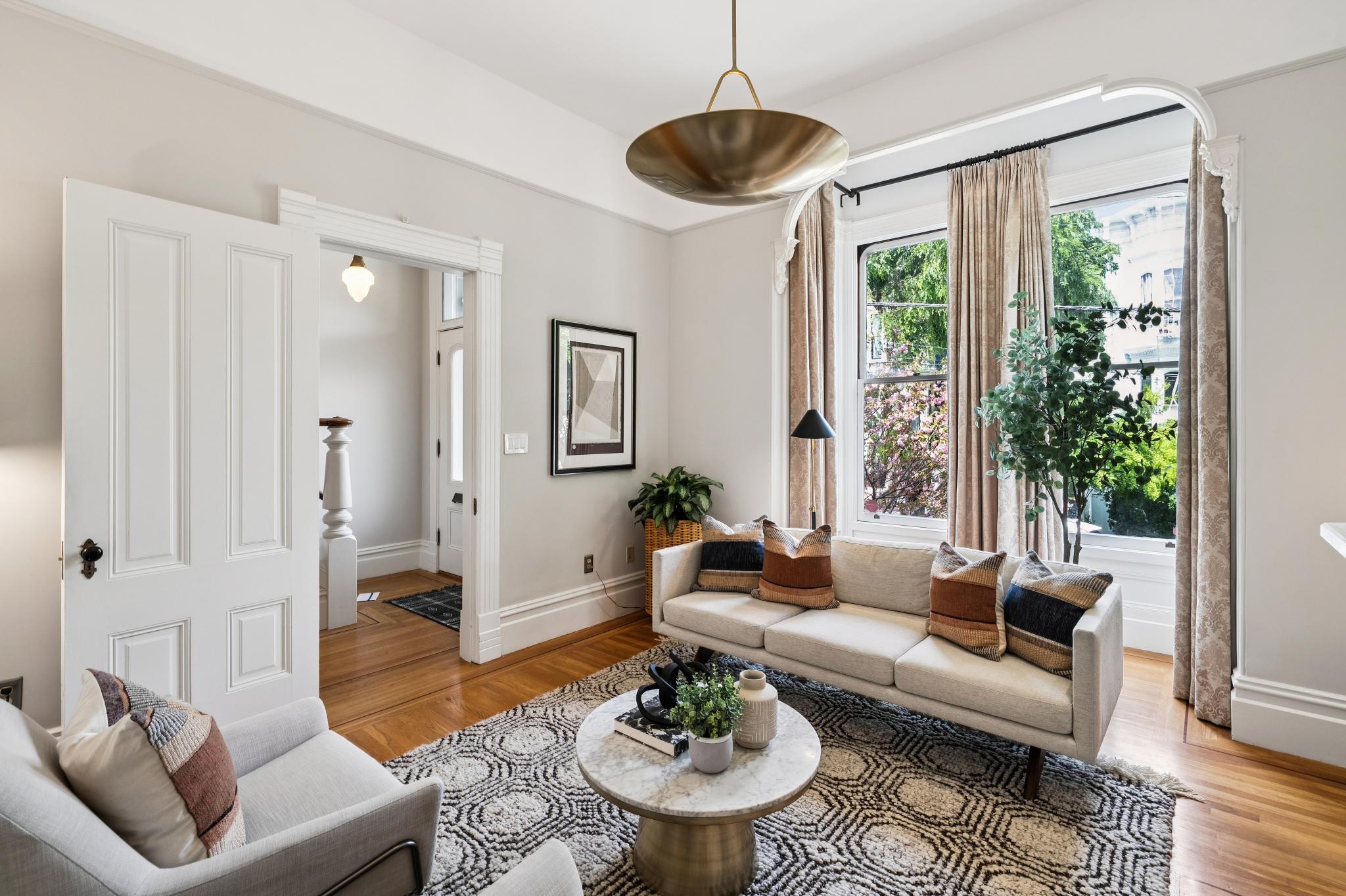 This bright and inviting living room features a modern neutral color palette, highlighted by a sleek light-gray sofa adorned with textured throw pillows and a sophisticated marble-topped coffee table. The space is anchored by a patterned area rug over warm hardwood floors, while a large brass pendant light serves as a striking focal point. Large windows draped in elegant curtains allow for ample natural light, creating a warm and airy atmosphere perfect for a contemporary home.