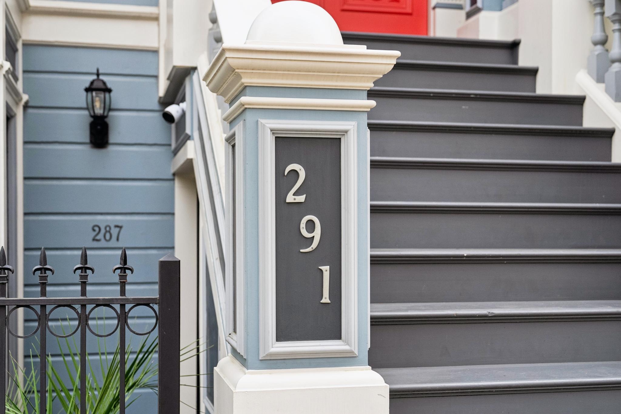 This close-up shot captures the elegant entryway of a classic home, featuring a prominent pillar displaying the house number '291' in crisp, white lettering. To the left, a glimpse of a neighboring property's '287' address is visible behind a decorative iron fence, while a set of dark grey stairs leads up to a vibrant red door. The architectural details, including the light blue trim and classic molding, evoke a charming, historic urban aesthetic.