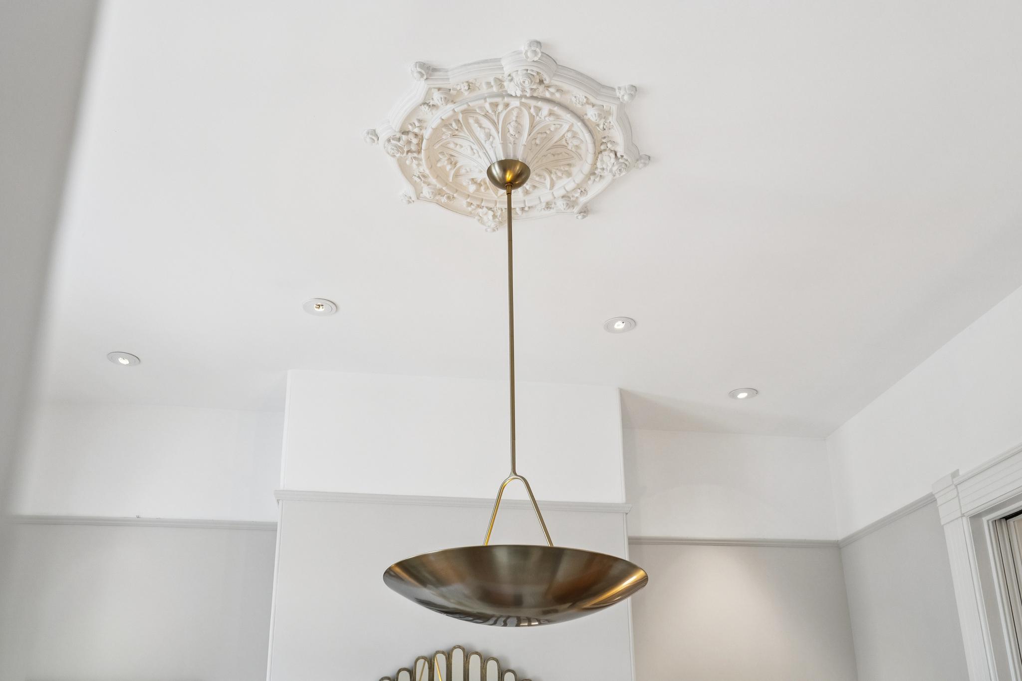 This image captures a sophisticated interior detail, showcasing an ornate, white plaster ceiling medallion that contrasts beautifully with a modern, minimalist brass pendant light fixture. The composition highlights the blend of classical architectural craftsmanship and contemporary design elements. The perspective is looking directly upward, emphasizing the height and elegant detailing of the ceiling.