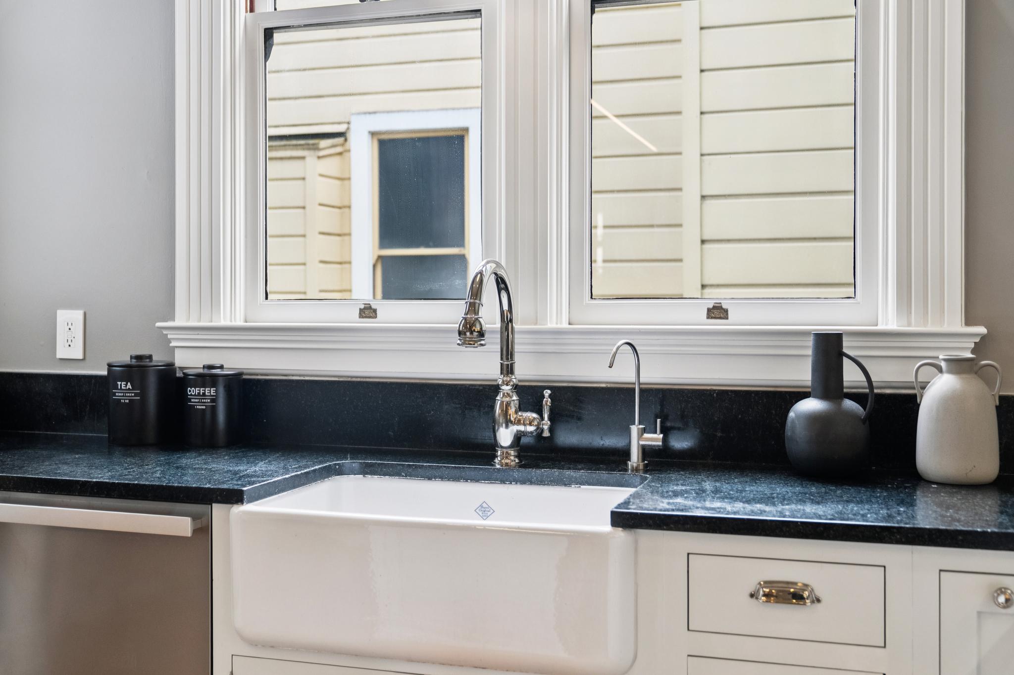 This kitchen features a classic farmhouse-style white apron-front sink set into a dark, polished stone countertop. A polished chrome faucet and a secondary water filtration tap are centered beneath a large, multi-pane window that brings in natural light. The space is accented by matte black canisters and minimalist decor, creating a clean, timeless aesthetic.