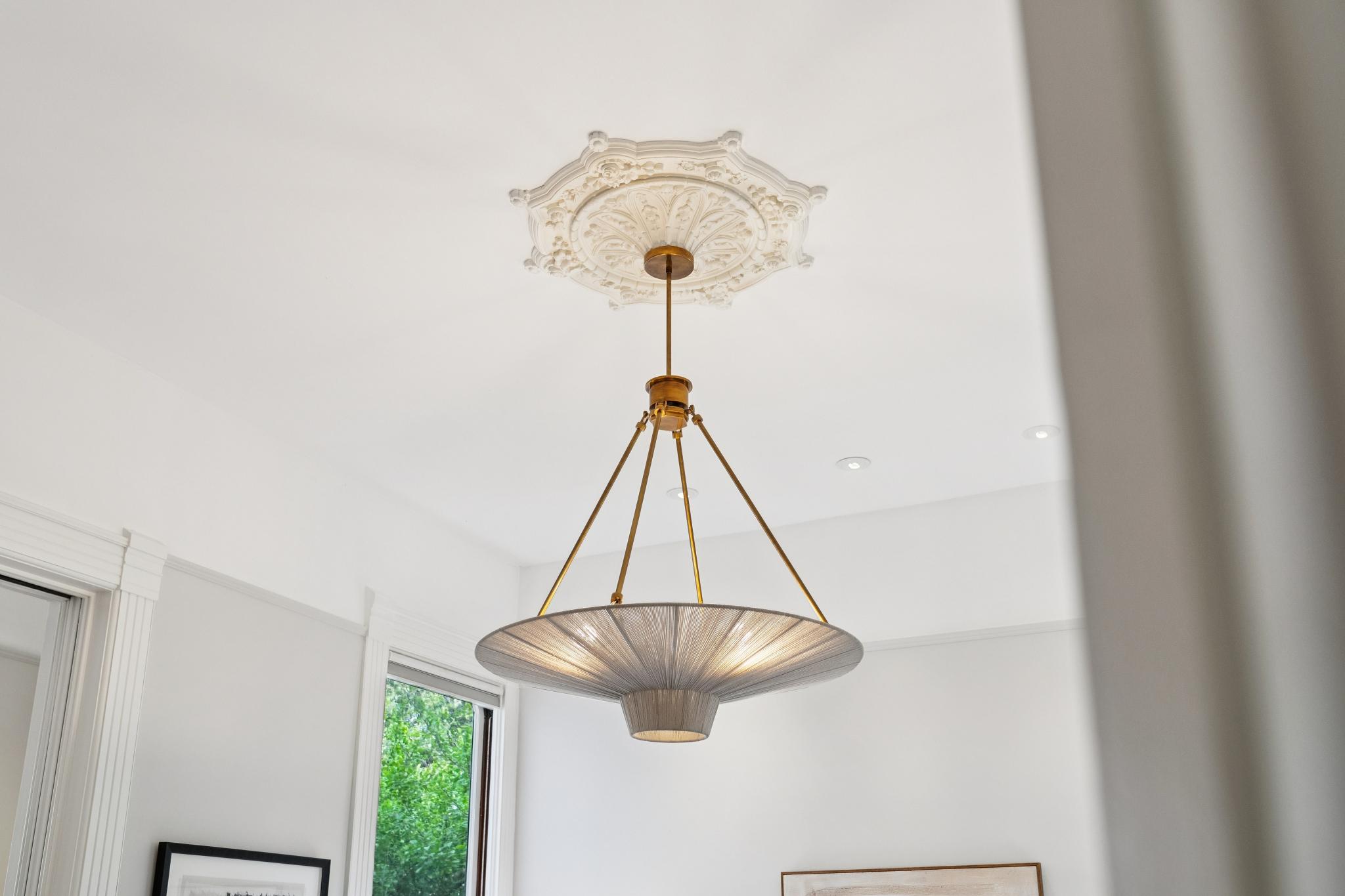 This image captures a sophisticated, modern pendant light fixture suspended from a ceiling adorned with an ornate, traditional plaster medallion. The contrast between the intricate, classic architectural detail and the sleek, contemporary design of the light fixture creates a refined and elegant aesthetic. The perspective is a low-angle shot looking upward, emphasizing the height and architectural character of the room.
