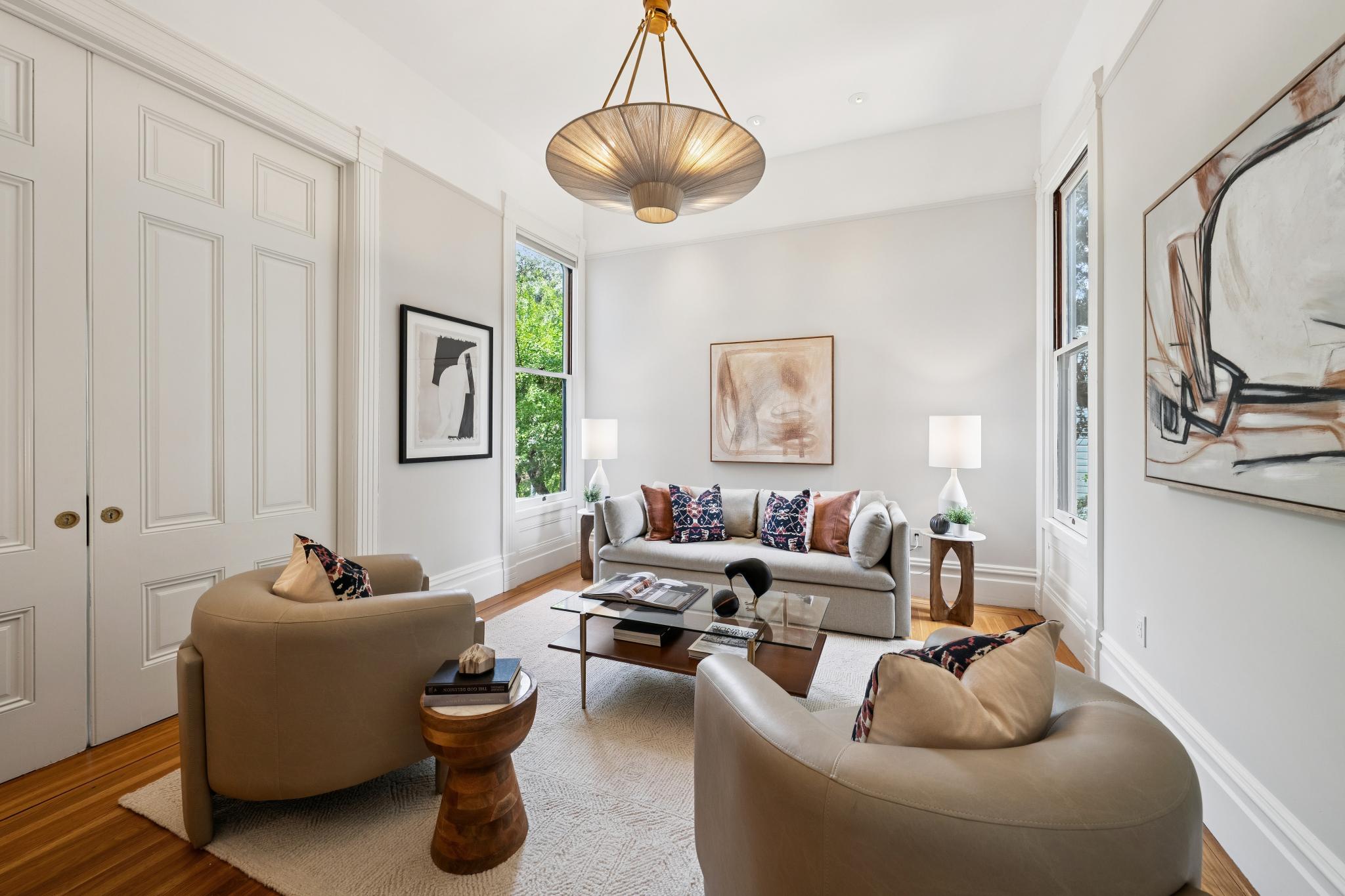 This elegant living room features a sophisticated neutral color palette, anchored by a plush light gray sofa and two curved leather armchairs. The space is highlighted by a striking modern pendant light, a glass-topped coffee table, and curated abstract artwork on the walls, creating a refined and inviting atmosphere. Large windows allow natural light to fill the room, emphasizing the warm hardwood floors and high ceilings.