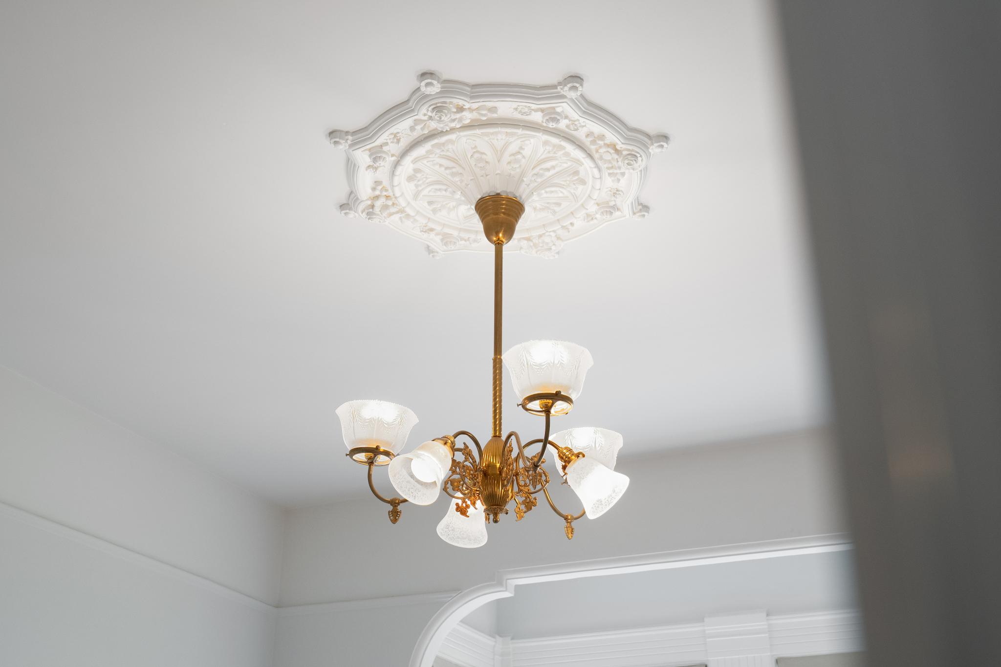 This image captures an elegant, vintage-style brass chandelier suspended from a highly detailed, ornate white ceiling medallion. The fixture features multiple frosted glass shades that cast a warm, inviting glow, highlighting the intricate craftsmanship of the metalwork. The perspective is a low-angle shot looking directly up, emphasizing the architectural charm and sophisticated character of the room's ceiling design.
