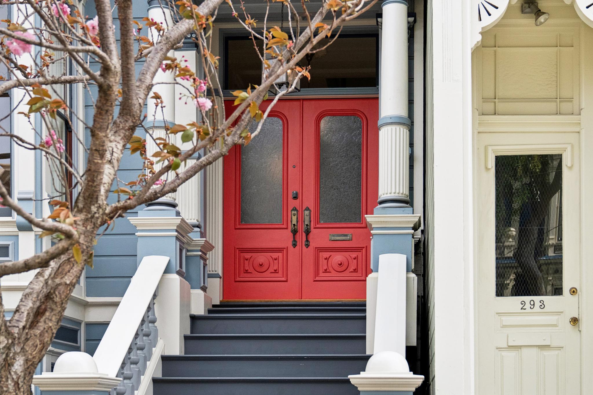 This image captures the charming front entryway of a classic Victorian-style home, featuring a striking set of double red doors with glass panels. The entrance is elevated by a set of dark-painted stairs, flanked by ornate white columns and blue-toned siding, while a blooming cherry blossom tree adds a soft, natural frame to the scene. The composition offers a welcoming and historic aesthetic, highlighting the architectural character and curb appeal of the property.