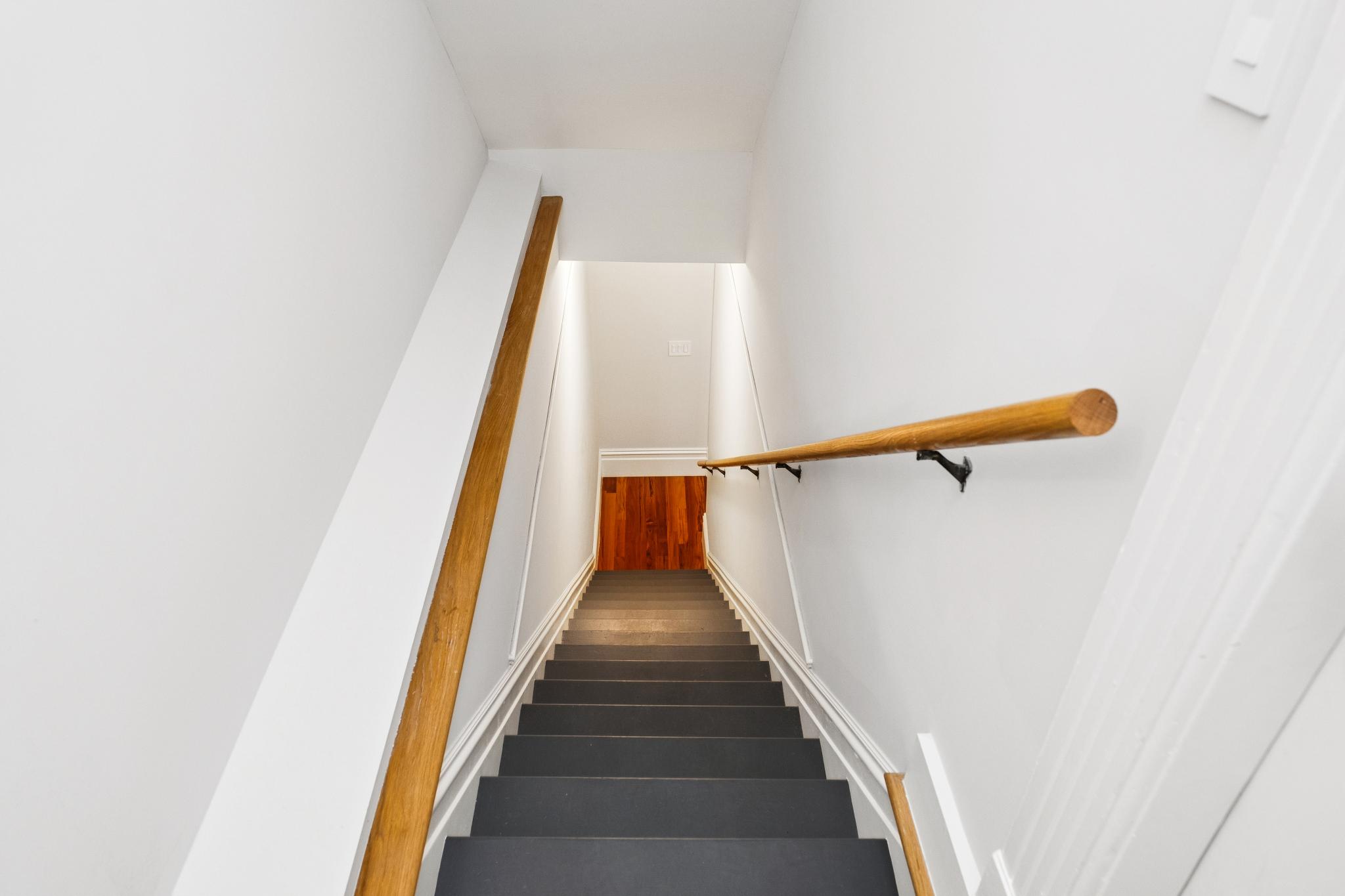 This high-angle, downward-looking shot captures a modern staircase featuring dark grey treads and crisp white walls. A natural wood handrail runs along the right side, complemented by a matching wood accent strip on the left wall, leading the eye toward a warm, hardwood-floored landing at the bottom. The clean lines and minimalist aesthetic create a bright, contemporary transition space within the home.