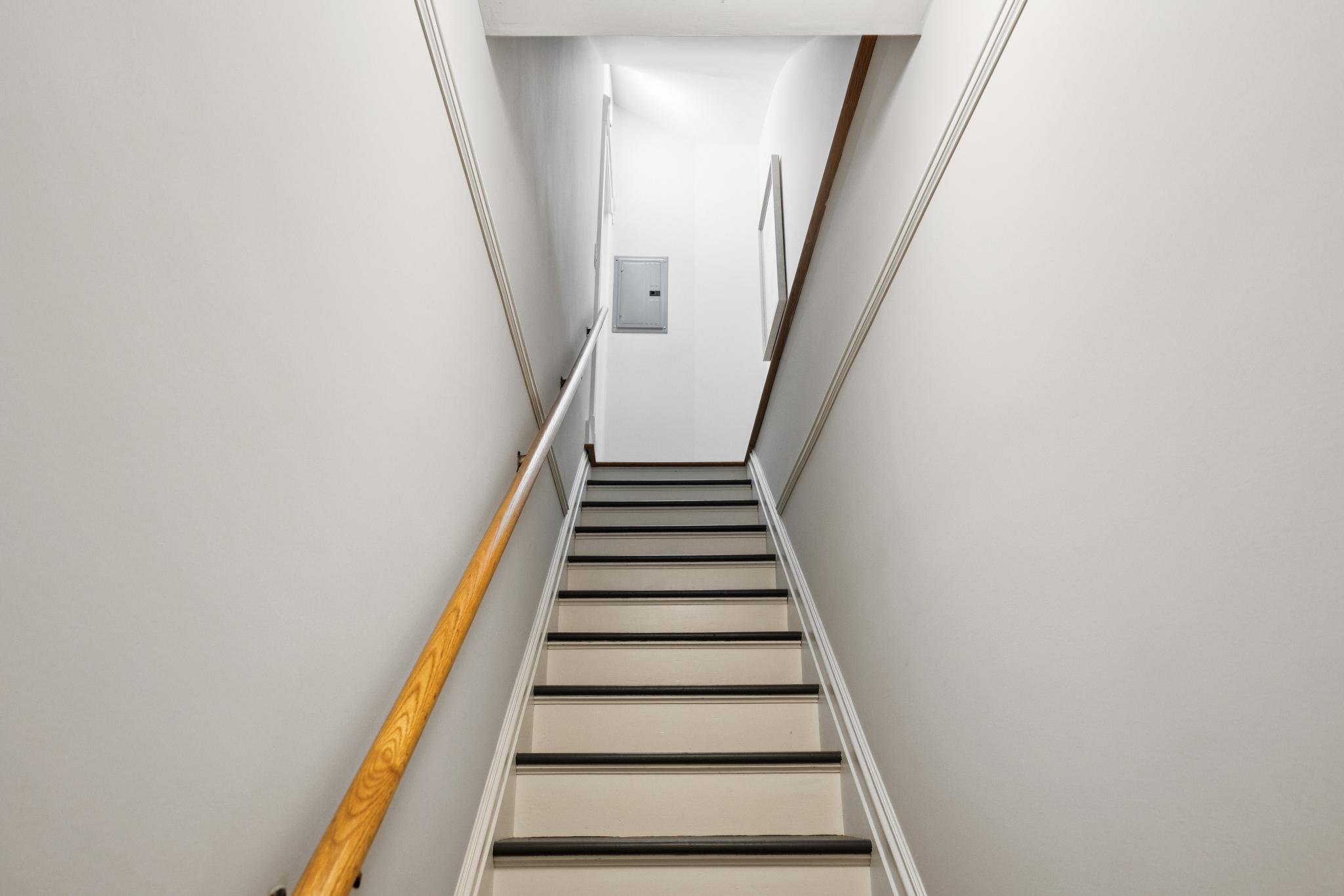 This image captures a straight, upward-facing perspective of a staircase featuring light-colored treads with dark non-slip edges. A wooden handrail runs along the left wall, which is painted a neutral off-white, while a small electrical panel is visible on the landing at the top. The composition is clean and symmetrical, emphasizing the architectural lines of the staircase leading to an illuminated upper level.