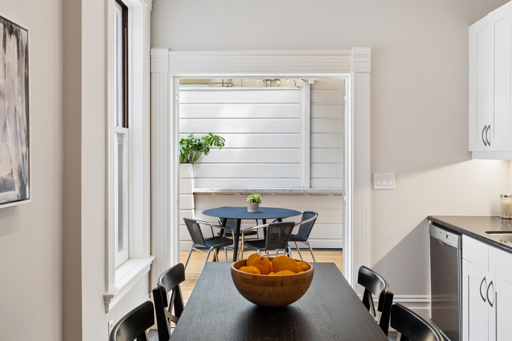 This dining area features a dark wood table topped with a bowl of fresh oranges, set against a bright, modern kitchen with white cabinetry. A large, open doorway leads directly to an outdoor deck, creating a seamless transition between indoor and outdoor living spaces. The deck is furnished with a round table and chairs, offering a cozy spot for al fresco dining.
