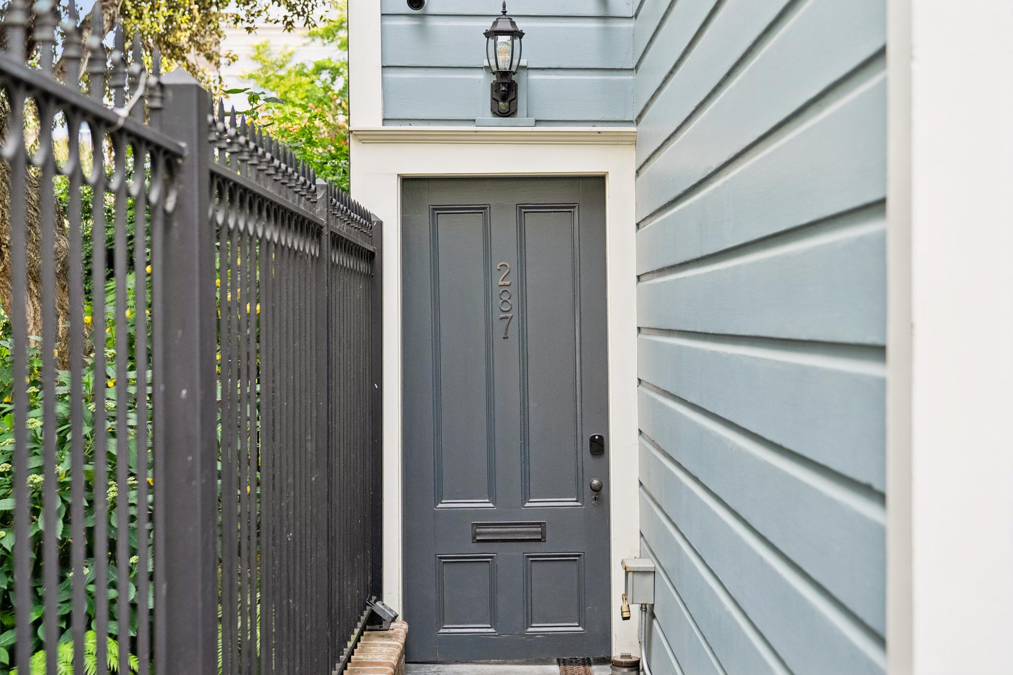 This image captures a charming, narrow side entryway featuring a dark grey paneled door marked with the number 287. The entrance is framed by light blue horizontal siding and a classic black wall-mounted lantern, creating a clean and inviting aesthetic. To the left, a tall black iron fence provides a sense of privacy, while the overall composition offers a focused, eye-level perspective of a well-maintained residential access point.