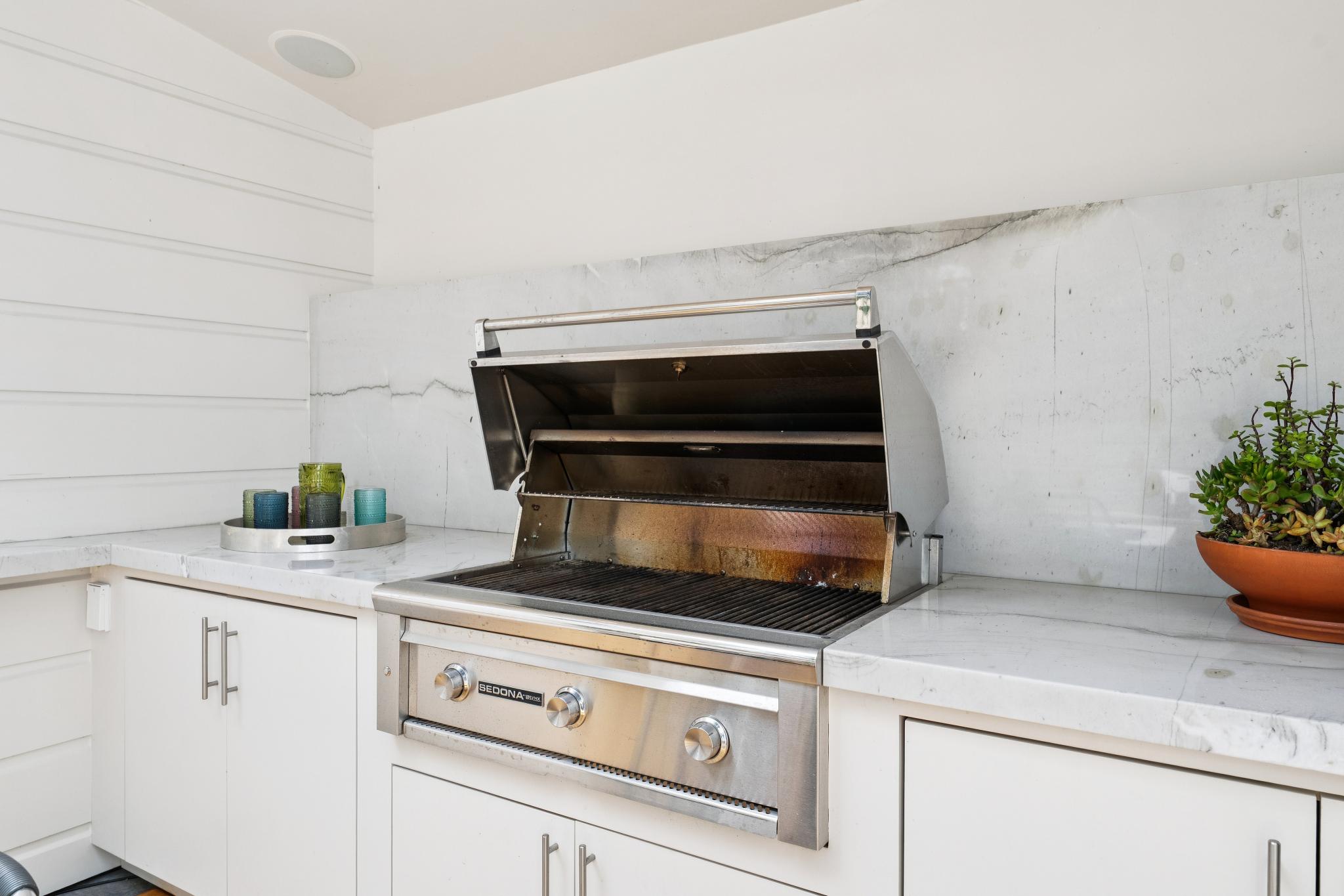 This outdoor kitchen area features a high-end stainless steel Sedona grill set into a sleek white marble countertop with matching backsplash. The space is accented by white shiplap walls and a potted succulent, creating a clean, modern, and inviting atmosphere for outdoor entertaining. The perspective is a straight-on eye-level shot, highlighting the premium finishes and functional design of the grilling station.