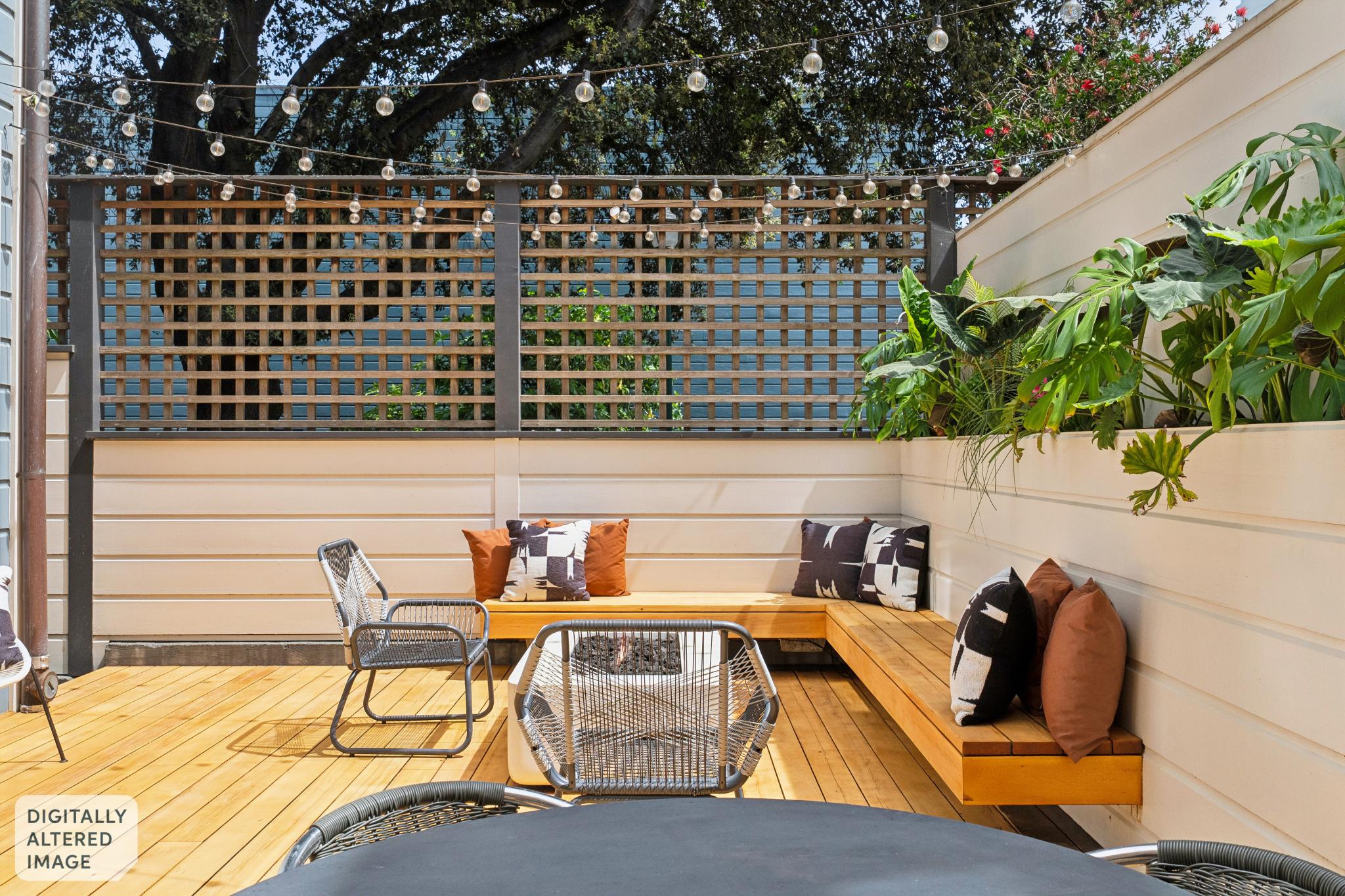 This inviting outdoor patio features a warm wooden deck with built-in bench seating arranged in an L-shape around a central fire pit. The space is decorated with stylish patterned and solid-colored throw pillows, complemented by lush greenery in a raised planter and string lights overhead. The scene is framed by a wooden lattice privacy screen, creating a cozy and modern atmosphere perfect for entertaining.