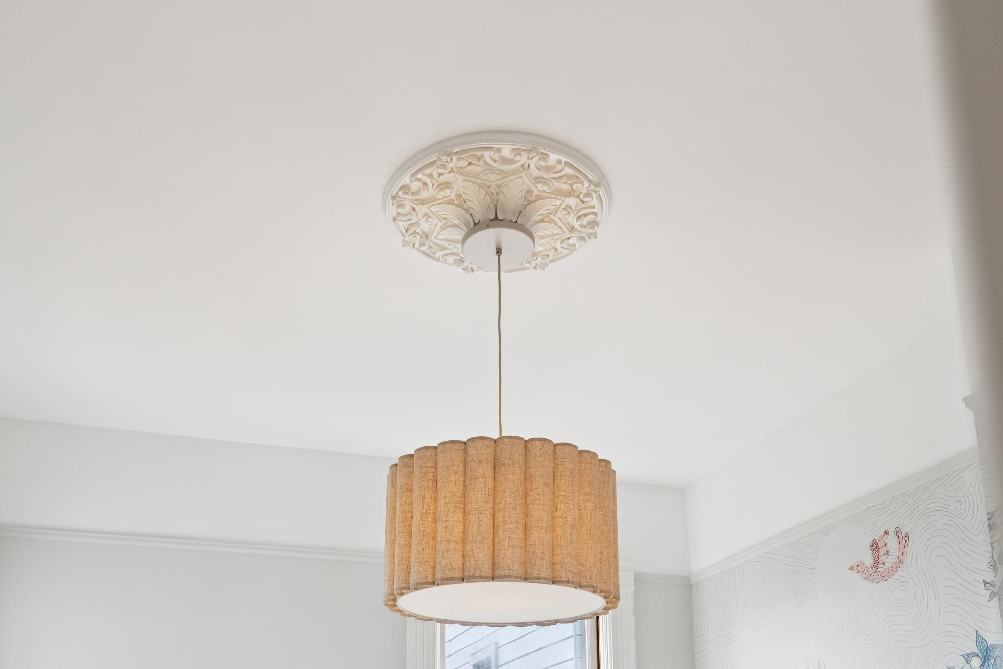 This image captures a stylish pendant light fixture suspended from a ceiling adorned with an ornate, white decorative plaster medallion. The light features a textured, pleated fabric drum shade in a warm beige tone, providing a soft, diffused glow. The perspective is a low-angle shot looking directly up at the fixture, highlighting the contrast between the intricate architectural detail of the ceiling and the modern, clean lines of the lamp.