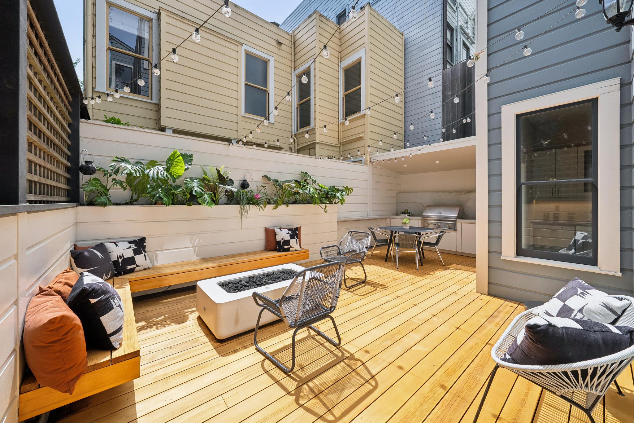 This inviting outdoor deck features a modern, multi-level design with a built-in wooden bench, a sleek fire pit, and comfortable contemporary seating. The space is enhanced by a raised planter box filled with lush greenery, string lights overhead, and a dedicated outdoor kitchen area with a grill. The warm wood tones and clean lines create a sophisticated, cozy atmosphere perfect for entertaining.