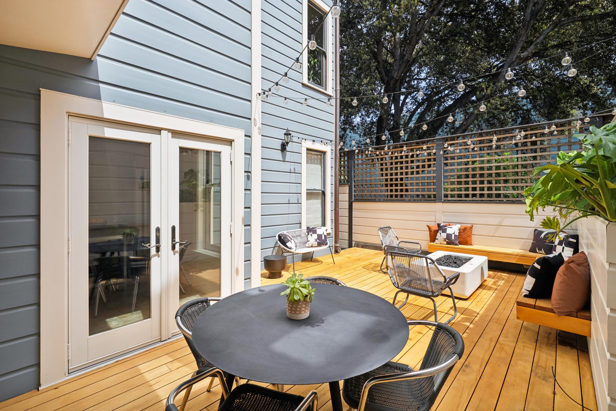This inviting outdoor deck features a modern, multi-functional layout with a dining area in the foreground and a cozy lounge space with a fire pit in the back. The space is framed by light blue siding, a wooden privacy lattice, and warm-toned wood decking, all illuminated by charming string lights. The overall aesthetic is contemporary and perfect for entertaining, offering a seamless transition from the indoor living area through the double glass doors.
