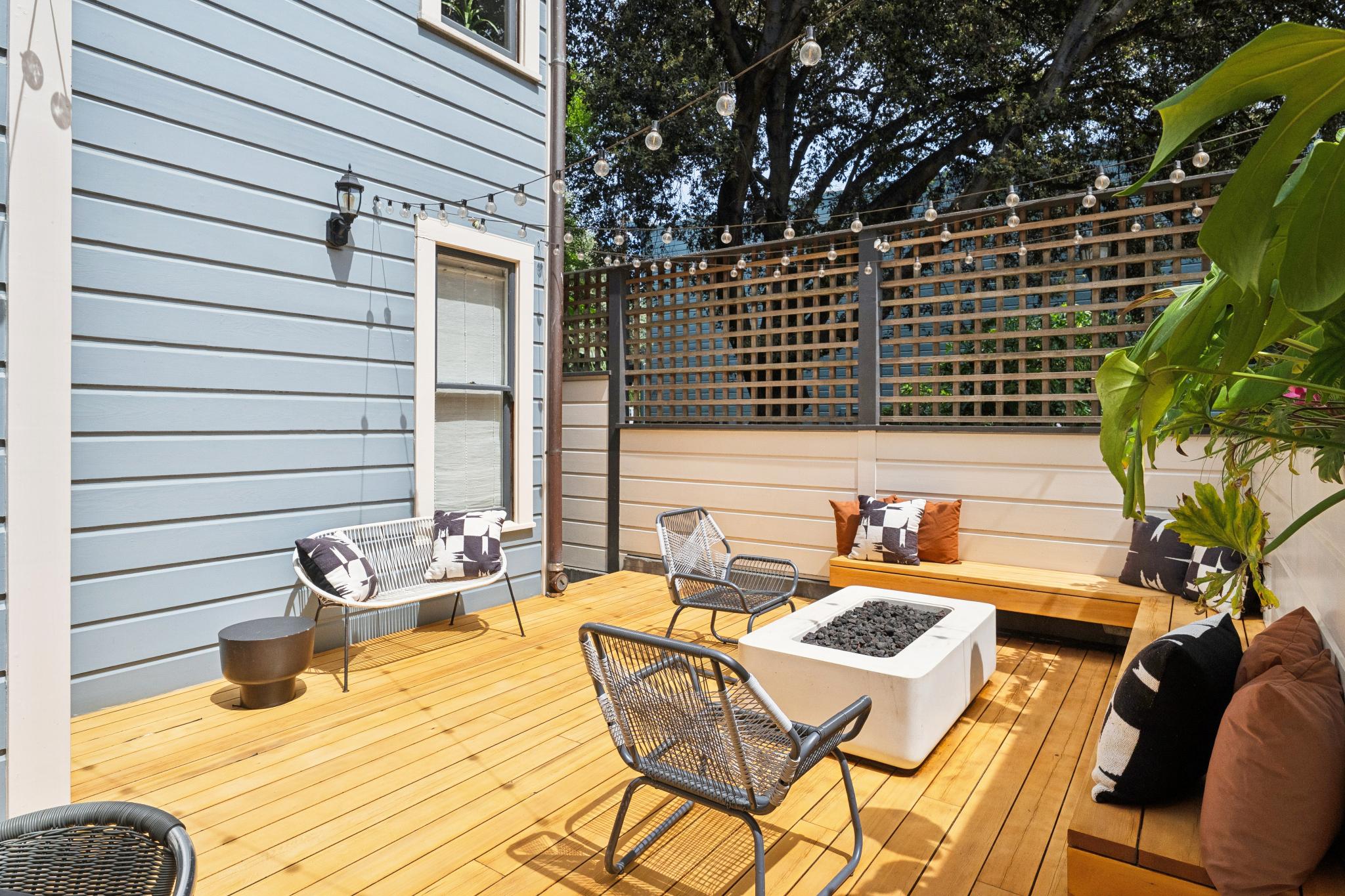 This inviting outdoor deck features a modern, light-wood finish and is furnished with stylish wire-frame chairs and a central concrete fire pit. The space is framed by a decorative lattice privacy screen and string lights, creating a cozy, intimate atmosphere perfect for evening relaxation. The perspective captures the entire seating area from a slightly elevated angle, highlighting the seamless blend of contemporary design and comfortable outdoor living.