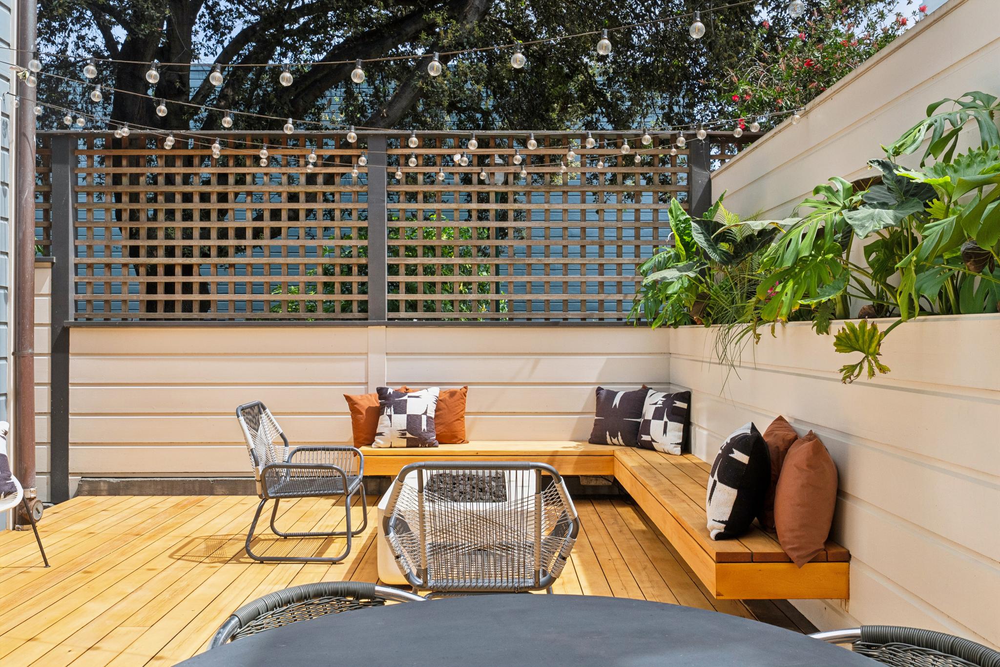 This inviting outdoor deck features a modern built-in wooden bench seating area arranged around a central fire pit, perfect for entertaining. The space is accented with stylish patterned throw pillows, lush potted greenery, and overhead string lights that create a warm, cozy atmosphere. The clean lines of the white horizontal siding and wooden lattice fence provide a contemporary, private aesthetic for this urban oasis.