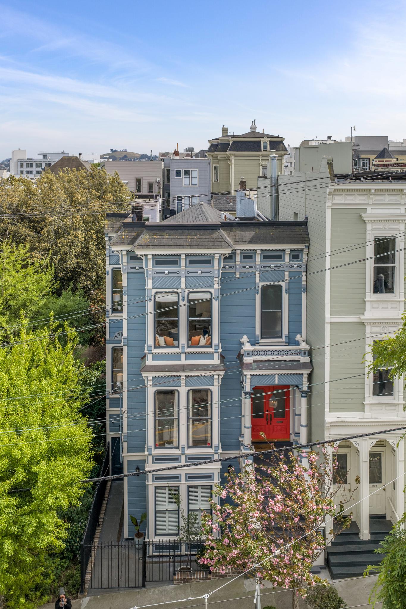 This image captures a classic San Francisco Victorian-style townhouse, characterized by its vibrant blue siding, intricate white trim, and a striking red front door. The multi-story structure features large, symmetrical windows and a charming, elevated entryway, all set against a clear blue sky. The perspective is a slightly elevated street-level view, showcasing the home's architectural detail and its integration into the dense urban neighborhood.