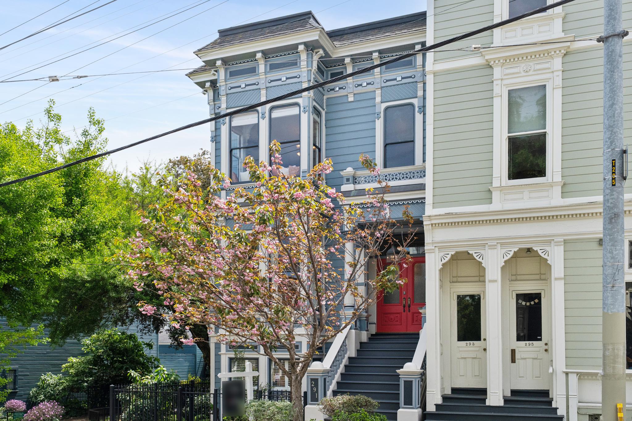 This image captures the charming front facade of a classic Victorian-style townhouse in San Francisco, featuring intricate architectural details and a vibrant blue paint scheme. A blooming cherry blossom tree stands prominently in the foreground, framing the bold red front door and the elegant staircase leading to the entrance. The composition offers a cinematic, eye-level perspective that highlights the historic character and curb appeal of the property.