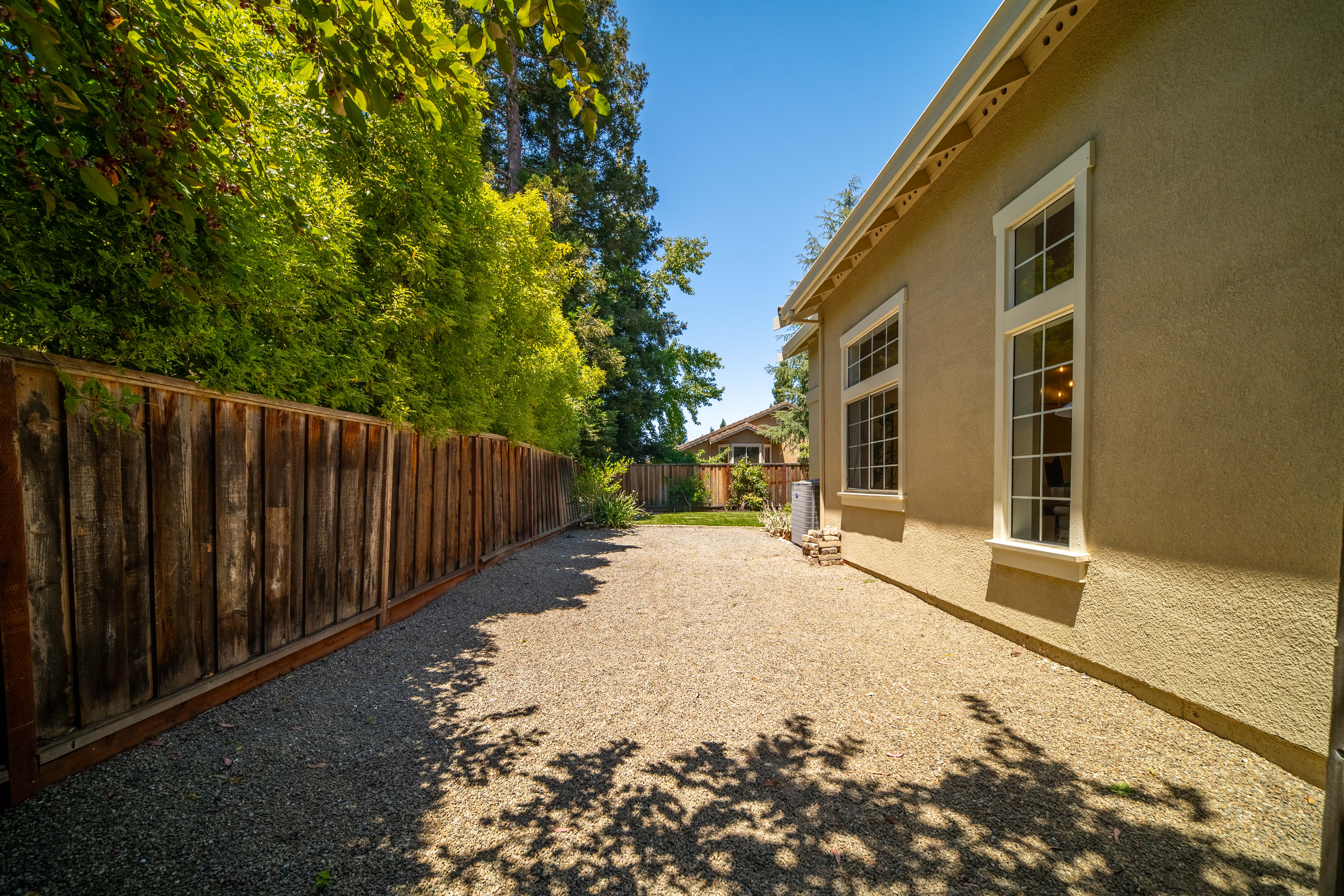 This side yard view features a long, gravel-covered pathway running alongside the beige stucco exterior of the home. A tall, weathered wooden fence provides privacy on the left, while large windows on the house allow for natural light. The scene is framed by lush green trees overhead, creating a quiet and private outdoor space.