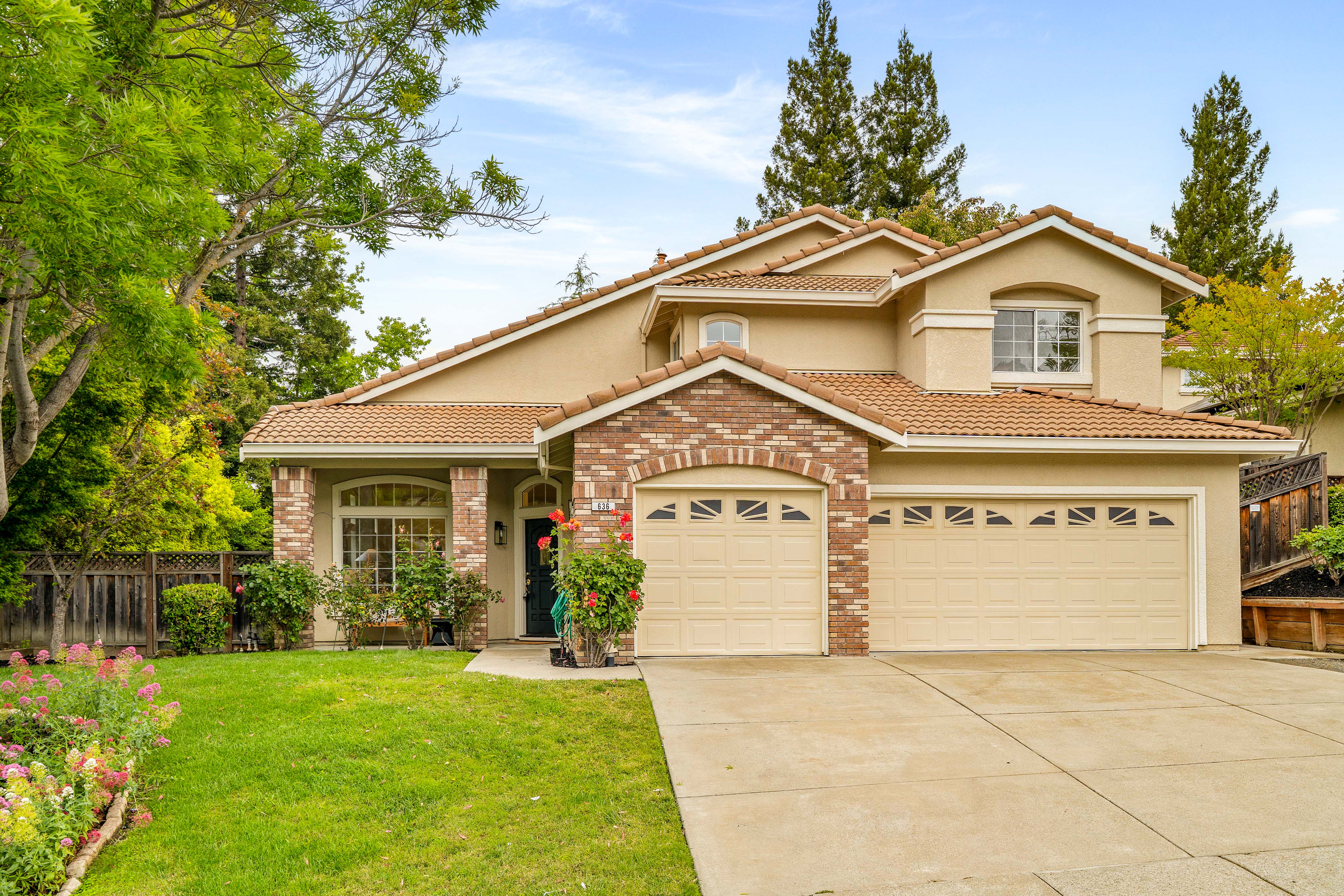 This two-story suburban home features a tan stucco facade accented with brick detailing around the garage and entryway. The property includes a multi-car garage, a well-manicured front lawn with vibrant flower beds, and a classic tile roof. The perspective is a straight-on eye-level shot, capturing the welcoming curb appeal and mature landscaping surrounding the residence.