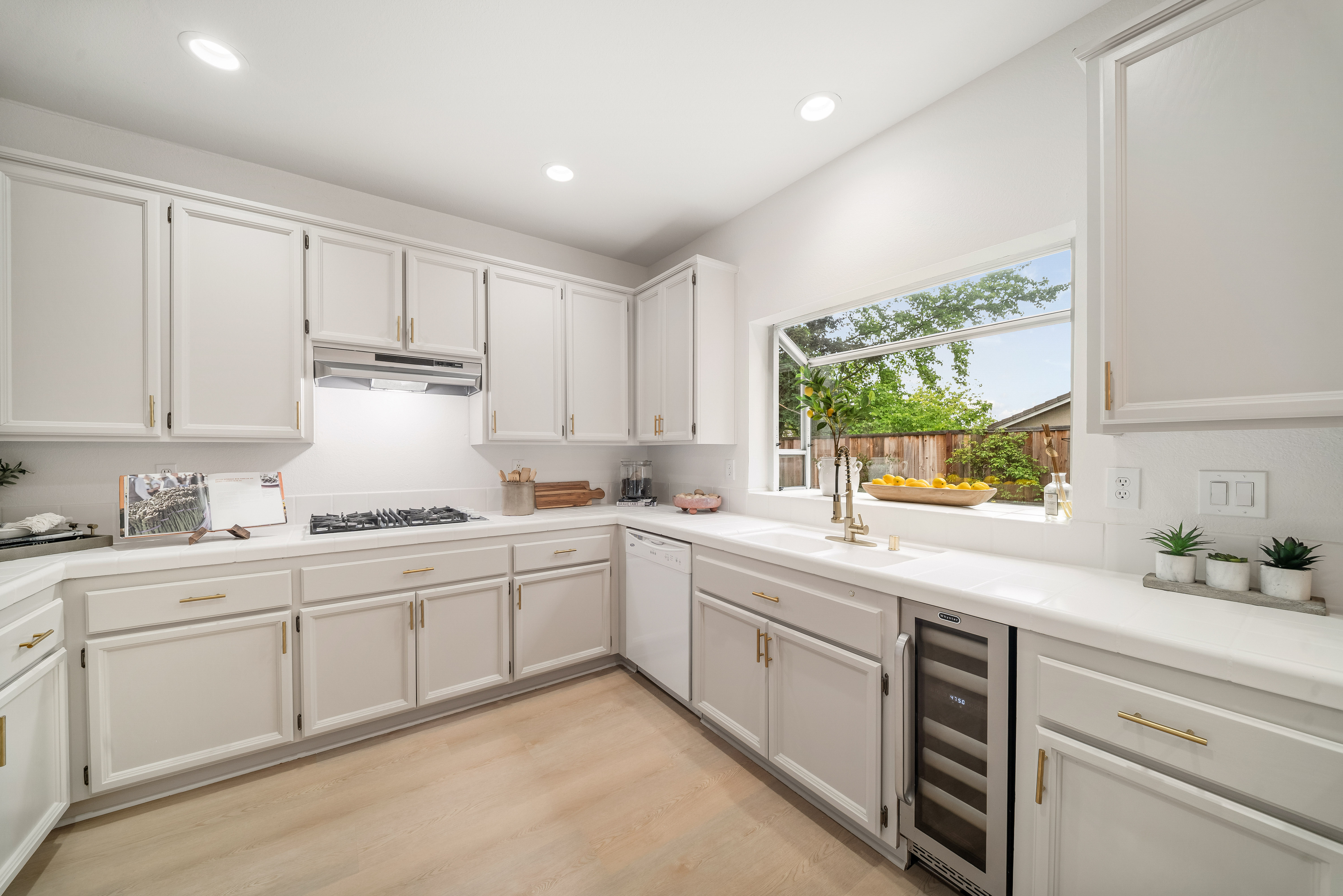 This bright and airy kitchen features crisp white cabinetry paired with light-toned countertops and matching light wood-look flooring. A large window above the sink provides a pleasant view of the outdoors, while modern gold-toned hardware adds a touch of elegance to the space. The layout includes a gas cooktop, a built-in wine cooler, and recessed lighting, creating a clean and functional culinary environment.