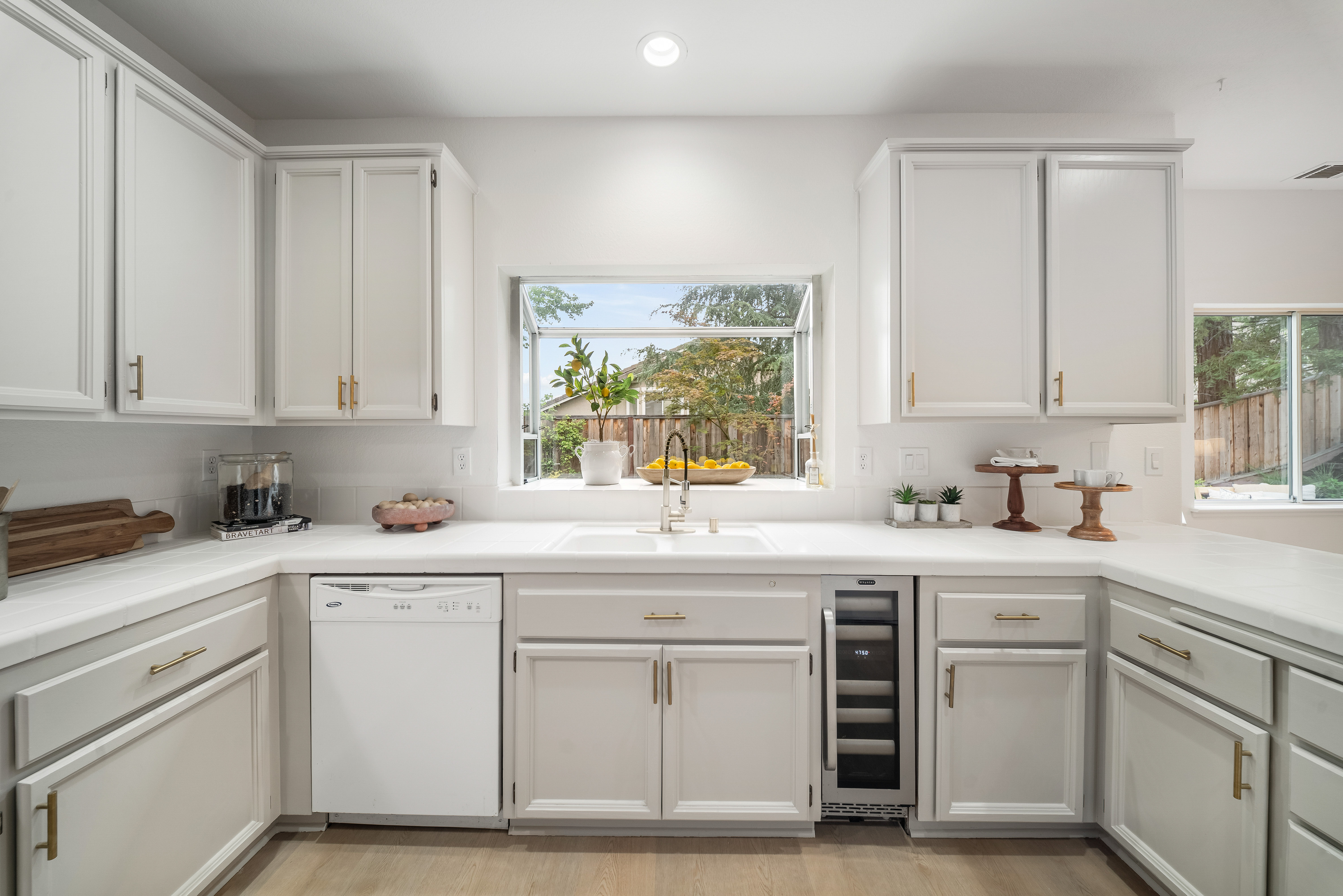This bright and airy kitchen features crisp white cabinetry with gold-toned hardware and a clean, white tiled countertop. A central window above the sink provides a view of the outdoors, while a built-in wine cooler and dishwasher are seamlessly integrated into the lower cabinetry. The space is finished with light-toned wood flooring, creating a welcoming and functional culinary environment.