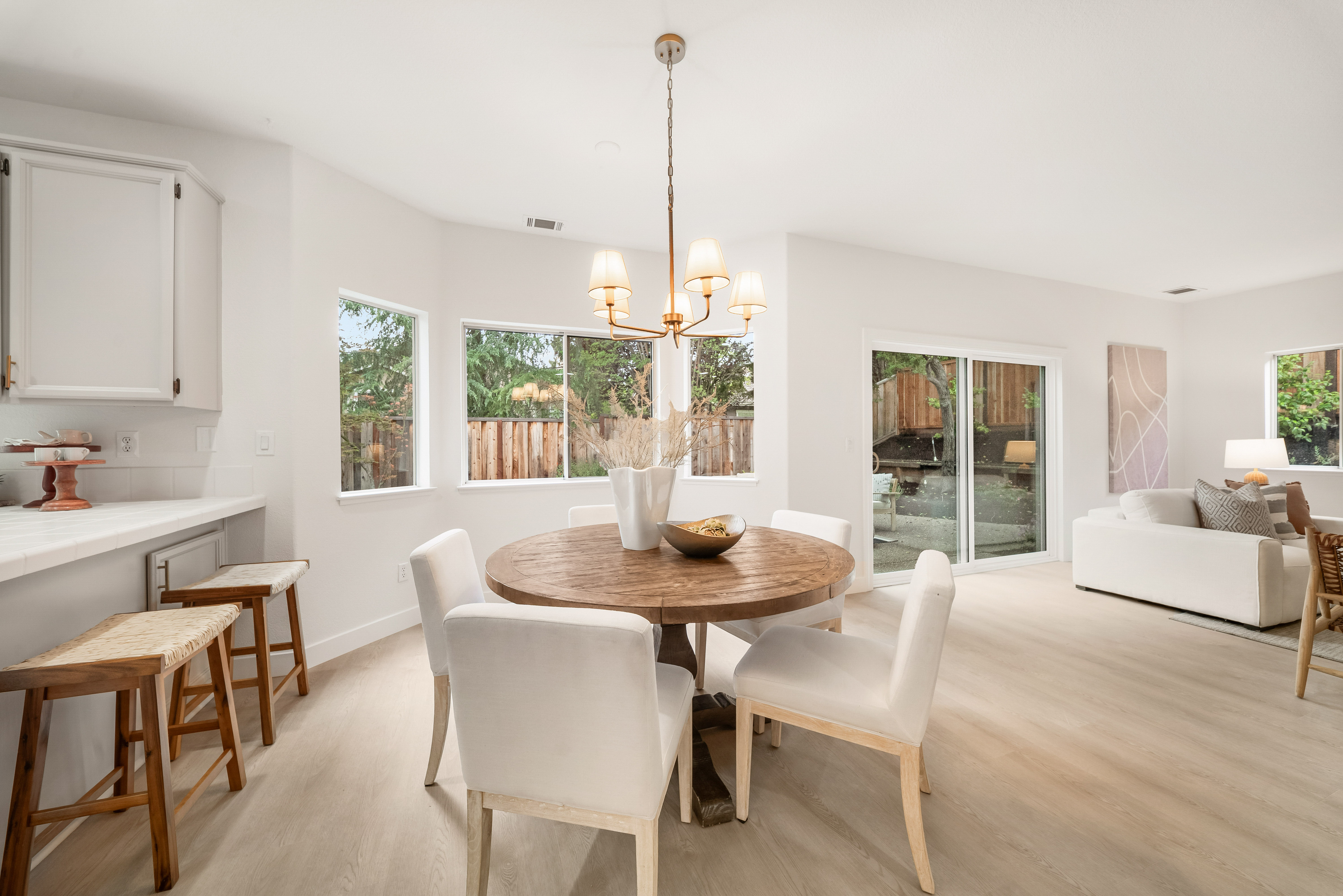 This bright and open dining area features a round wooden table surrounded by four upholstered chairs, positioned beneath a classic chandelier. The space flows seamlessly into a living area with light wood-toned flooring and large sliding glass doors that provide a view of the backyard. A breakfast bar with two stools connects the dining space to the adjacent kitchen, creating an airy and cohesive open-concept layout.